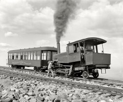 Pikes Peak, Colorado, circa 1900. "Summit, cog wheel train, Manitou and Pike's Peak Railway." 8x10 glass negative by William Henry Jackson. View full size.
How they did it.Taken a few years ago:
Dinosaurs in the mountainsThe sister to this locomotive, M&amp;PP #4, is still nominally operable and is as far as anyone knows the only operable Vauclain compound. The two cylinders you see are both power cylinders; the valves are hidden on the inside. Vauclain compounds, named after the head of Baldwin, were briefly popular around 1900 but fell out of favor along with most other compound locos with the introduction of the superheater; in this case part of the problem was unequal forces from the two pistons which produced wear problems at the crosshead. On this little bitty engine it apparently wasn't too bad a problem.
Nice Trip!Today a ticket on the Railway costs $34. You can hike up and take the train down, but if you miss the last train and have to be evacuated, the fee per hiker is $500!
I'd stick with the train. Looks lovely.
http://cograilway.com/Pikes%20Peak%20train%20videos-A.htm
Oh My. Call a Tow Truck, er Train"But officer, just look -- that passenger car was heading the wrong way on my side of the tracks. Now how am I ever gonna get the front end of my engine out from under it??"
Cog and Pinion Appliances


Crofutt's Overland Guide, 1892. 

The Manitou &amp; Pike's Peak Railway, a recent organization, commences at a point just above the Iron Springs and runs to the summit of Pike's Peak. The road is about 8&frac34; miles in length. The average grade is 18 per cent.,the maximum being 25 per cent. and the minimum 8 per cent., with 16 degrees curvature. The rails are the standard T rail, with a double cog-rail in the center, weighing 110 tons to the mile. Each engine has three cog and pinion appliances, which can be worked together or independently; in each cog appliance is a double set of pinion brakes that work in the cog, either of which when used can stop the engine in 12 inches going either way, on any grade and at a maximum speed of eight miles an hour. Fare for "round trip," $5.00.



The Street Railway Journal, April, 1893. 


Manitou &amp; Pike's Peak Railroad,

which is known as the "Cog Wheel Railroad," and which runs to the top of Pike's Peak, a distance of about about 8,000 ft. higher than Manitou. The road was opened for traffic in October, 1890. The fare for the round trip is $5, and the round trip is made in about three hours. The rack, which is placed midway between the rails, consists of two steel bars, notched to a depth of about two and a half inches, with teeth staggered, and which are firmly fastened to the ties by means of bolts and shouldered chairs.
The engines are of peculiar shape, and the power is transmitted to two pinions located under the boiler, which mesh with the gear of the rack, so that sufficient power is obtained to force the engine and car up the steepest grades. Only one car trains are run, and the cars are pushed ahead of the engines in ascending, and return in the same relation. The engine and car are not coupled, but there are bumpers consisting of perpendicular and horizontal steel cylinders about five inches in diameter and eighteen inches long, which provide for the varying grades and angles. The car, as well as the engine, is equipped with pinions which mesh into the rack and which are controlled by powerful band brakes, so that the car can be controlled independent of the engine, every known safety appliance being employed to prevent the possibility of an accident.
Formerly, high pressure engines were employed, but during the last season one compound engine was run, and the other three engines have recently been sent to the Baldwin Locomotive Works where they are being made over into compounds. The line is operated only during the summer months, as the accumulation of snow upon the mountains during the winter prevents the running of the cars.



Journal of the Association of Engineering Societies, 1894.


Manitou and Pike's Peak Railway.
The Engines

During construction and the first year's operation, the Pike's Peak Railway had three engines built by the Baldwin Locomotive Works. These weighed about 26 tons each, loaded with fuel and water. The cabs and boilers of these engines were much like those of ordinary locomotives, but here the resemblance ceased, for the bearing-frame of the engine was inclined, so that the boiler was level on a 16 per cent. grade, the average grade of the road. The engine had no tender, water being carried in two tanks at the side of the boiler, and coal in a box at the rear of the cab, holding one ton. The engine rested on three axles, the forward two being rigidly fastened to the frame, while the rear one was furnished with a radius bar, the rigid wheel-base being 6 feet 8 inches, and the total wheel-base 11 feet 2 inches. To the two forward axles was fastened an inside frame carrying three sets of two pinions each, making six pinions in all. The specifications for these pinions called for hammered crucible steel, with ultimate tensile strength of 100,000 pounds per square inch, stretch 16 per cent, in 8 inches, the teeth to be cutout of the solid disk.

SynchronicityWhat a coincidence! We just rode the Pikes Peak Cog Railway three days ago with children and grandchildren. It still takes about three hours, and it is an amazing ride. Temps were about 90 degrees in Manitou Springs and below 50 degrees at the summit. We even had a little skiff of snow up top. We passed by the original water cranes that supplied the early steam engines. The trip is spectacular, but I kind of wish I could have taken it in the steam days. (Of course, I would be dead by now, right?) It was great to get back to internet civilization and find this picture on Shorpy!
(The Gallery, DPC, Railroads, W.H. Jackson)