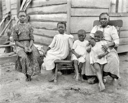 Circa 1902, location not specified. "A happy family." 8x10 inch dry plate glass negative, Detroit Publishing Company. View full size.
Spitting imageThe girl second from the left looks like a clone of her grandmother!
What Grandma is sayingNobody knows the troubles I've seen. Looks like they've had a rough life. Happiness is innocence.
HappyI've been staring at this picture for ten minutes, now. At first, I just thought it was a very sad picture, but I am seeing more in it, now.  The expressions on their faces don't look like they were miserable, despite how truly dirt poor they were. Maybe the pleasant looks on their faces came from the fact that they possessed that super-human quality of valuing loved ones so highly that they felt thankful, despite having little else but each other. The girl on the left looks like she is starting to blossom into a beautiful young woman. Her posture is very elegant and lady-like. The grandmother's hands have obviously worked very hard, quite certainly from the time she was a small child. 
I hope their economic situation improved at some point. They have really touched my heart and I will remember them. 
I felt sorry because I had no shoesWhen I see such a photograph, two things always come to mind:
1) the good old days were frequently overwhelmed by primitive living conditions and considerable poverty; and
2) we certainly take a lot for granted.
What were their dreams and aspirations? What impact did poverty have upon their daily lives? Did they ever obtain relief?
The older woman would have known the institution of slavery firsthand. How did she describe that to the younger children?
Count your blessings.
SatchThe little boy sitting in his grandmother's lap will be a jazz musician in the 1920's!
Quote from King Solomon"A merry heart maketh a cheerful countenance."  The kids do appear happy and acknowledged.  Unconditional love has a way of causing that.  Material riches don't really enter into that equation but a strong, caring relative might.
Not enough informationThey don't tell us whether Grandma's watching the youngers while the parents work, or if this is all the family that there is.
But that dust looks wonderful for smooshing bare toes around.
(The Gallery, DPC, Kids)
