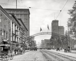 Columbus, Ohio, circa 1910. "High Street north from State." 8x10 inch dry plate glass negative, Detroit Publishing Company. View full size.
+94Same view from July of 2009.
ArchesWere those arches really only for street lighting and flags?
CuriousWhat is that tall building on the right; it looks very modern, even in the early photo.  Anyone know?
Arch CityColumbus was called the Arch City for the dozens of arches spanning its streets. The first were wood, put up in the 1890s. Then replaced by metal. The last of them seem to have been removed by the mid-teens.
[Reproduction arches are recent additions to some streets.  - Dave]
Open All NightThank you, Google, for finding that old football program (a must-see pdf!) in the back pocket of my old suit trousers.
As it turns out, Leachman's Chop House is at 61 S. High Street, and Mykrantz Drugs (the name just faintly has the right letterforms on one sign) is just a few doors to the north.
At the far right of the frame is the McKinley Memorial monument, built 1906.  I'll be sitting on the bench there with a newspaper and a cigar for a little while, before getting on with my day.
Early RadioI wonder if they had an early wireless station there at that time? In the upper left of the photo, there is a vertical tower atop the building and what appears to be insulators in the guy wires. This is a normal practice in a live vertical radio frequency radiator (antenna) so that the guy wires do not become resonant at the operating frequency, as well as insulating the antenna from ground.
A bit early for broadcasting but two-way code wireless telegraphy was done all the time by then.
[The appearance is indeed similar to wireless masts of the era. - Dave]
The View In 1914Here's a picture of roughly the same view dated as 1914. Notice how many more cars and fewer horses there are in the picture.
Leachman&#039;s Chop HouseI'd love to stop by Leachman's Chop House for lunch (infamous in a web search for being where Ohio State's Sphinx Senior Honorary club was born), saunter next door to Bryce's for some suits, hats and shoes, pick up a nice ten-cent cigar at the drugstore (so frustrating! can't quite make out the name!), sit in the park across the street and contemplate what goods require the services of the "Press Post" specialty pressing parlor!
But what I'm really here for is to find out what is being offered at 4%, or 4% off, as per the giant numbers on the top of the building just ahead on the right!
p.s. It's a little hard to sort out all the wires and metal posts and arches, but it looks like the arches were put up for decorative (and perhaps useful) lighting on alternating sets of the metal poles that hold up the trolley wires.
62 South HighThe address here is about 62 S. High Street. At the far right you can just make out the McKinley Memorial, dedicated in 1906.
(The Gallery, DPC, Streetcars)