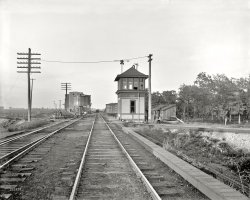 Circa 1900. "Chicago &amp; Alton Railroad. Signal station and crossroads at Mazonia, Illinois." 8x10 inch glass negative, Detroit Publishing Co. View full size.
Iron Mountain BabyThe boxcar behind the tower is from the St Louis, Iron Mountain and Southern Railway, which is famous in railway folklore for the Iron Mountain Baby.
Route 66About 26 years later Route 66 would follow alongside the northern Illinois part of the Chicago &amp; Alton RR from Chicago down to Springfield.
Do RR folks know why the wires are running so close to the ground on the right?  I assume the covered raceway covers the mechanical links to switches behind the photographer.  
MZThe "MZ" sign designates the telegraph code for this interlocking tower. Each operating point on the railroad had a unique code assigned to it. 
Re: Iron Mountain BabyWhat a great story! Thanks for the link!
The photos and meticulous work restoring and adjusting them make this site a wonderful resource, but the comments often enhance the stories immensely. Just one more thing I love about Shorpy!
No PlatesWhat wonderful detail of track construction at the turn of the century.  This photo still shows that they were not using tie plates (metal plates placed between the crosstie and rail to make the structure more stable and keep the rail from cutting into the ties).  Amazingly, other than the rail becoming heavier and larger through the years, most of the other features of construction remain to this day.  Main lines have mostly gone to continuous welded rail these days, eliminating those "clickity-clack" joints, but industrial and older yard tracks still have the angle bars (splice bars) and bolts joining each length of rail. There also has been little improvement in the design of those crossing frogs, where the steel wheels still must bang across about an inch gap in the rail used to accommodate the flanges of the rails for the crossing route.
Cable conduitI've only been in one railroad switching tower in my life, and that was nearly 50 years ago, but I'm pretty sure the switches were all mechanically operated. So those "wires" along the track are probably the cables running from the operating levers (you can just see the tops of them in the tower's windows) to the switches. 
Flag Stop?There's no town here, and probably never was one; that mass of trees conceals a great expanse of marsh. Lacking my 1948 Handy Railroad Atlas I couldn't tell you whose line cut across the C&amp;A here, but in any case the track is gone. If you look at the Google aerial view you can just barely find a trace of it to the northwest. There's still a junction here, though: the line that bends off to the left (see the semaphore to the left of the handcar) is still there.
The wires down on the ground most likely controlled the signals; it looks a bit haphazard but I don't think there's enough room under the boards to put rods for everything.
Gandy Dancersfixing the track, and those cables on the left are to operate switches down the track, in the foreground you can see where the cables go under the track to the opposite track.
(The Gallery, DPC, Railroads)
