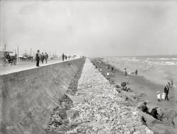 Circa 1905. "Seawall and beach at Galveston, Texas." 6½ x 8½ inch dry plate glass negative, Detroit Publishing Company. View full size.
Galveston Seawall historyThis is probably the first section of the seawall [construction began on this section in 1902 and was completed in 1904], which was built as a result of the Galveston Hurricane of 1900. This initial section was a little over 3 miles long, and would be extended over the years as the Island's population grew westward.
Probably the most interesting thing about the construction of the seawall was the grade raising that was also done as part of the seawall project. The bulidings, homes, and utilities on the east end of the Island [where the majority of population was located at the time] were raised as high as 17 feet, with the space below filled with mud and sand pumped from the floor of Galveston Bay--quite an engineering feat for its time. Check this web site for detailed information and photos concerning the grade raising: 
http://www.therealgalveston.com/Grade-Raising.html
For over 100 years, the Seawall did exactly what it was intended to do--protect Galveston from major hurricane damage. Unfortunately, the Seawall met its match in Hurricane Ike in 2008, when the huge storm surge swept over the wall and caused massive flooding, even in the highest part of town directly behind the wall.
But, to show that the Island residents' sense of humor survived Ike, I was down there earlier this year and was in some bar on the Strand (old downtown district). A line was painted on the wall about 7 feet above floor level, indicating the "Hurricane Ike High Water Mark", and just under that, "You must be at least this tall to get drunk in this pub!"
Post-HurricaneIn 1900, a huge hurricane devastated Galveston, killing thousands. Part of the problem was the lack of a seawall. The highest point in the city was only 9 feet above sea level, so when the storm landed with its 15 foot tidal surge, the result was utter destruction. The seawall shown here was built in 1902, and is now 17 feet high. It stood up to the 12 foot storm surge of a 1915 hurricane, though Hurricane Ike in 2008 spilled water over it.
RebarThat has to be a pioneering use of reinforced concrete, yes?
Hurricane of 1900Just finished reading the book "Isaac's Storm" by Erik Larson about the extremely deadly hurricane that hit Galveston in 1900. According to Larson, this seawall was built as a result. About 8,000 people died, and it was costlier than Hurricane Katrina.
Off the mapThe seawall and grade raising were significant engineering feats of their time.  However, they couldn't stop the loss of prestige that Galveston suffered after the storm -- Galveston had been the most prosperous and glamorous city in Texas before the 1900 hurricane, but was later surpassed by Houston and Dallas.
Another good book to read is John Edward Weems' "A Weekend in September." Published in 1957, Weems interviewed numerous survivors who offered gripping, and sometimes inspiring, eyewitness accounts of the horrors of that weekend.  An interesting note is that one could still see high water marks from the flood on some of the since-raised houses, even in the 1950s.
Galveston Had a Seawall"Galveston had a seawall
To keep deep water out,
But the high tide from the
Ocean spread the water all about.
Wasn't that a Mighty Storm"
-- Huddie Ledbetter
Point of orderThe board of engineers that designed the Galveston seawall was chaired by retired Brigadier General Henry Martyn Robert (not Roberts), best remembered to posterity as the author of the Pocket Manual of Rules of Order for Deliberative Assemblies, more readily known as Robert's Rules of Order. 
Hurricane IkeA lot of Ike's damage in Galveston was the storm surge that came from the bay side of town, which is not protected like the beach side.  The historic commercial district The Strand took 8 feet or more of water.  I was down there for Mardi Gras 2009, and someone had painted an Ike "high water mark" on a building in The Strand.
The WallIt is my understanding (from being a local - living in an evac zone on the mainland just north of Galveston) that the seawall did its job during Ike, but that the flooding was from the storm surge moving around the seawall, especially through the harbor entrance and to a lesser extent, the non-protected west side. The surge inundated the island from behind. The good aspect of this is that it was flooded rather than scoured with a powerful tidal force, like Bolivar on the other side of the harbor with no sea wall. The storm surge fiercely destroyed, scoured and eroded the peninsula, while Galveston proper (behind the seawall) was more 'gently' flooded.
The seawall is still only going to serve mostly to break the forward advance, but the fact is the island slopes down and away from it toward the bay, and any significant storm surge will flood. The surge won't slam and scour, though.
Hurricane Ike was not too bad of a wind storm, its damage was the massive, unusually large surge. Wind-wise, Galveston has endured worse, we even kept our roof, but surge-wise it was massive! Without the seawall it would have been cataclysmic. Good engineering 100 years old still doing its job!
Looking eastThis may have been taken around 15th or 16th Street looking east, as you can see where the wall makes a jog to the left, out of sight, in the distance. That's between 12th and 13th Streets.
Other serious hurricanes came through in 1909 and 1915, and it was discovered that the small rocks shown here at the foot of the wall were insufficient to protect it, and larger, pink granite "rip-rap" were put down instead.
In the distance one cane see a steam crane, possibly at work laying the rocks at the foot of the wall farther east.
(The Gallery, DPC, Swimming)