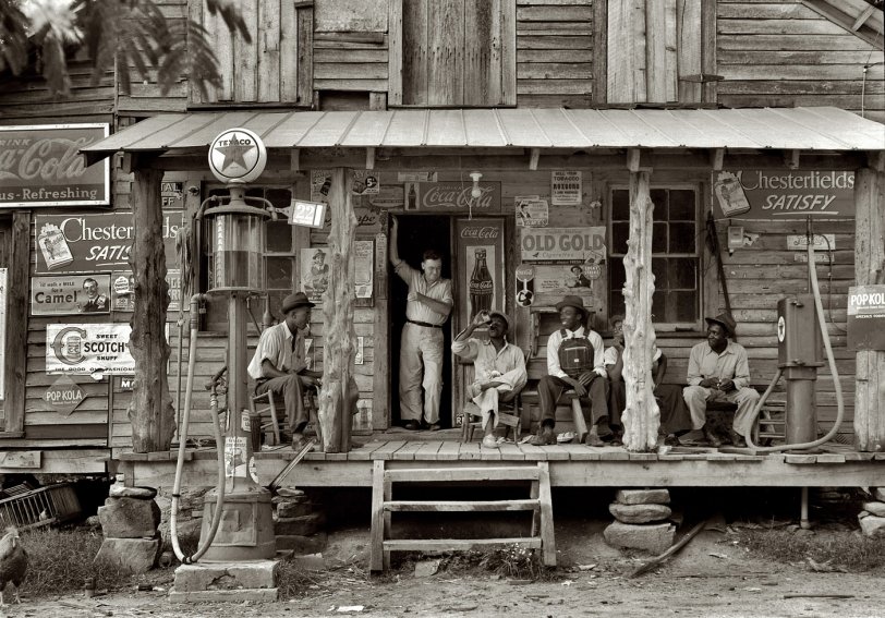 July 1939. Gordonton, N.C. "Country store on dirt road. Sunday afternoon. Note kerosene pump on the right and the gasoline pump on the left. Rough, unfinished timber posts have been used as supports for porch roof. Negro men sitting on the porch. Brother of store owner stands in doorway." 4x5 nitrate negative by Dorothea Lange for the Farm Security Admin. View full size.
