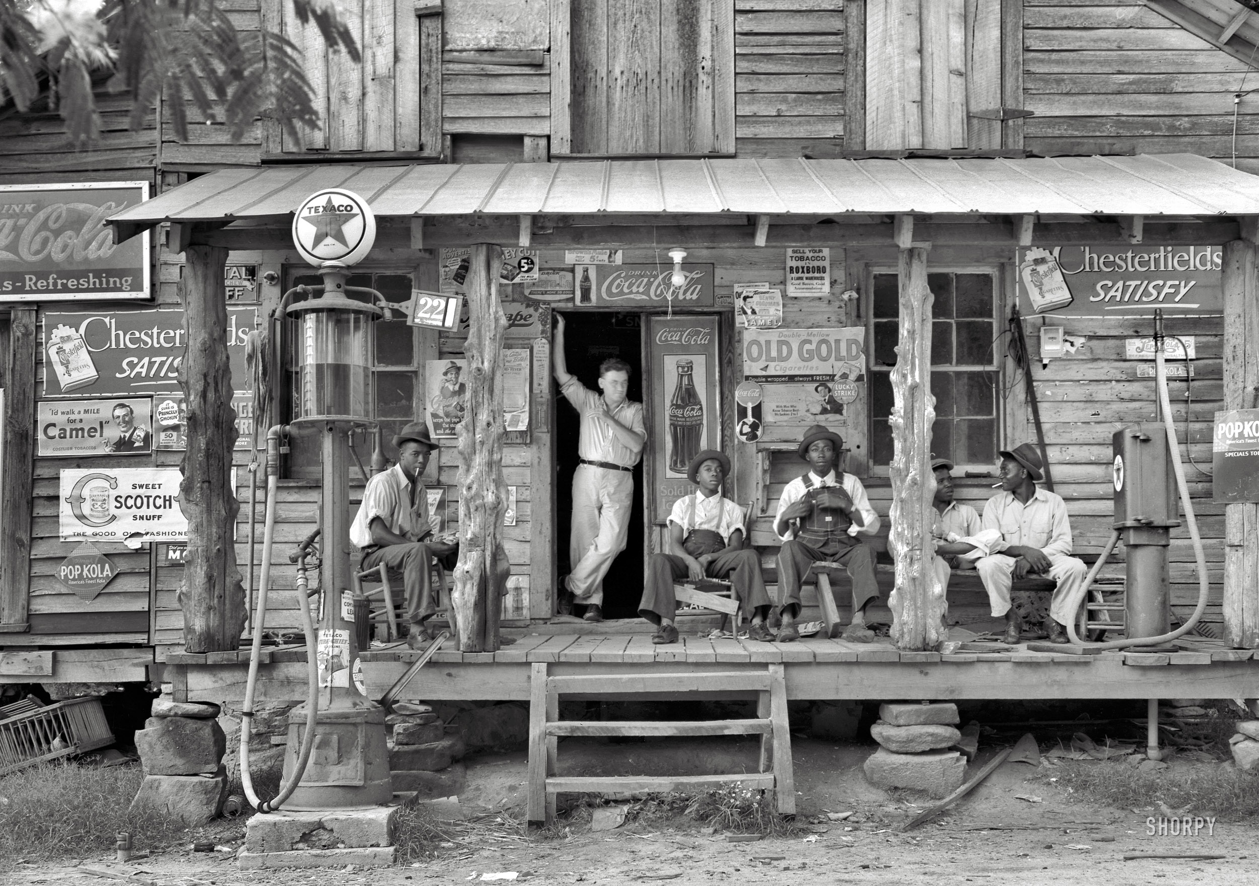 July 1939. Gordonton, North Carolina. "Country store on dirt road. Sunday afternoon. Note kerosene pump on the right and the gasoline pump on the left. Rough, unfinished timber posts have been used as supports for porch roof. Negro men sitting on the porch. Brother of store owner stands in doorway." Our second look at this establishment, seen here two years ago. 4x5 nitrate negative by Dorothea Lange for the Farm Security Administration. View full size.