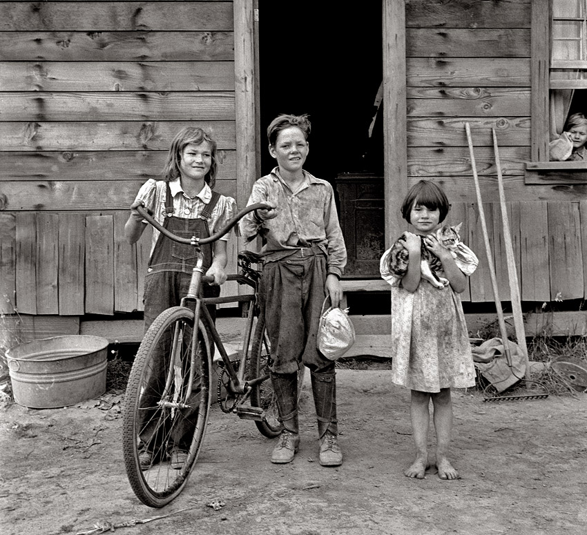 August 1939. Three of the four Arnold children outside their farmhouse at Michigan Hill. The oldest boy earned the money to buy his bicycle. Thurston County, western Washington. View full size. Photograph by Dorothea Lange.