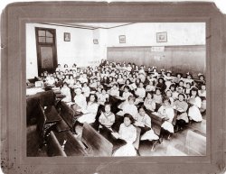 This picture of Anna Maggi (far left side of room) was taken around 1910 in an unidentified Catholic school in the old Borough Hall section of Brooklyn, N.Y. On the classroom blackboard is written "Month of the Holy Souls" which would be November. View full size.
Modern Urban EducatorI am an educator in an urban school.  My classes of 25 - 30 students are very challenging.  
There seems to be an assumption that schools were better "in the good old days" and modern educators are not up to the task.  Can one really assume that this classroom of children is better educated than a classroom of 25 today? Where all of these students expected to attend college or to pass a state mandated test?  Are these girls read for an job market that isn't even identified yet? It's time we stopped assuming that our education (or in this case, our grandparents' education) is superior to a modern education simply because it worked for us.
Class SizeThe class size seems to be similar to what I recall from Our Lady of Lourdes in Bethesda MD in the 50's - about 60 pupils. Today, "learning suffers" if class size exceeds 25, yet somehow we all emerged literate, numerate, and well disciplined. 
Why nuns needed rulersA quick rows-by-columns estimate counts 90 girls in that room!  I'd guess the standers in the back usually sat in the empty section on the right, but moved to get into camera range.
And we worry today about a class size of 25.
(ShorpyBlog, Member Gallery)