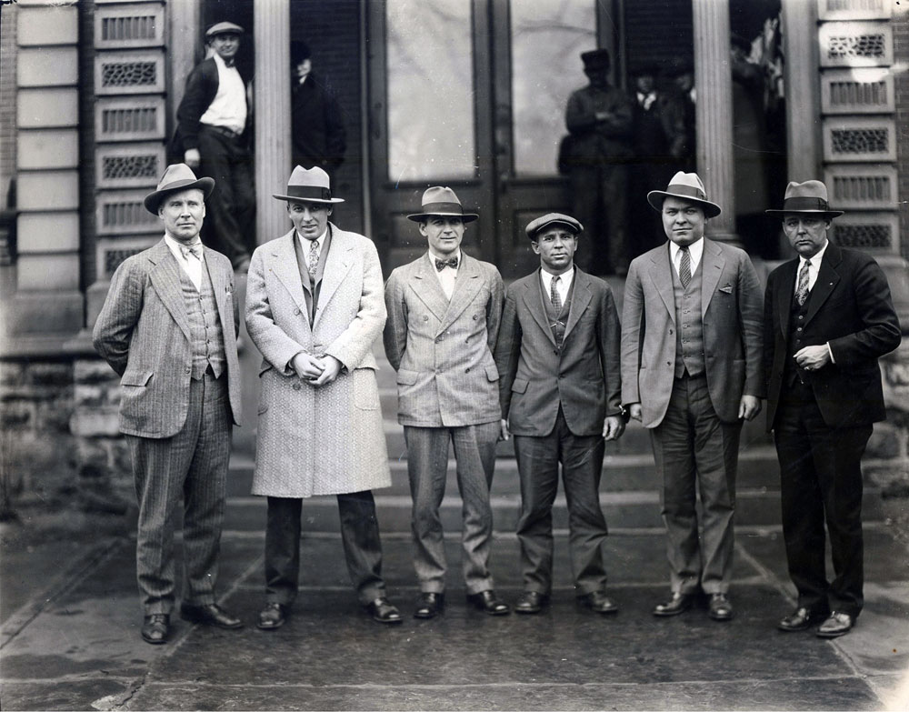 From back of photograph: "The four men suspected of robbing the Linngrove (Indiana) Bank, with Sheriff Hollingsworth (left), and Prosecutor Nathan Nelson (right), were caught by the Journal Gazette Photographer as they were leaving the Adams County Court House, Friday morning after being arraigned for the robbery. Between the sheriff and the prosecutor, reading left to right are: Evertt D. Gaither of Fort Wayne, J. Phillip Chamberlain of Columbia City, Elmer Wood of Pennville and Robert E. Siniff of Fort Wayne. Photograph appeared in the Journal Gazette on 3/23/1929. Photo by Clippinger." View full size.