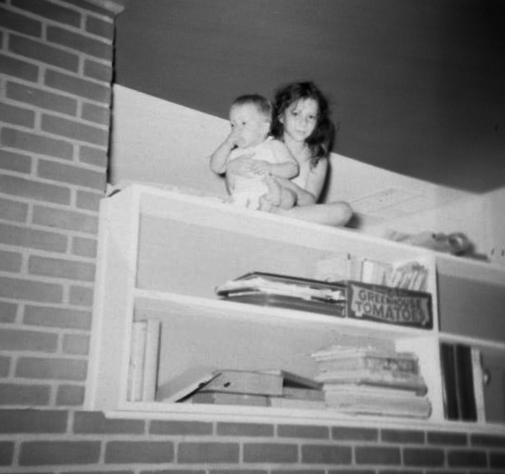 Eight-year-old me clutches my one-year-old brother in the space above the giant fireplace of our Dogwood Drive Levittown home. How I got up there is simple. I climbed using a combination of a kitchen step stool and those shelves you see below me. How I got my baby brother up there was probably my parents’ worst nightmare. My best guess is that I stood on the step stool and put him there before I climbed up.
 
The dark angle you see above my head is not a room corner. This shelf was open air on both sides. My mother had the ceiling of the white kitchen painted bright red. That is the edge of the kitchen ceiling paint transitioning to the white of the living room ceiling. The kitchen/living room wall is across the right center of the frame. This was at the very end of the Levittown era of my childhood. Our next home in Princeton, New Jersey was being built for us by this time.

Since one of my parents took the picture, they clearly found out what I had done. I suspect they found out after I had done it, not before. I was not punished in any way, but they might have suggested that I play up by the ceiling myself, and leave my living doll brother for more floor-based hiding places and games.