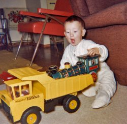 Here I am, Christmas 1964 with my new Tonka dump truck.  I vividly remember sitting in the dump bed and riding it down our sloped driveway, bending the bed sides outward. View full size.
Hard bedfellowsMy brother, born in 1959, got one of those for Christmas, probably the year before that.  I remember that he insisted on having it in bed with him, at night, alongside his Teddy bear! The bear's name wasn't Teddy, though, it was "Joses." That was his version of Bill Dana's character from the Ed Sullivan Show, the "astronaut" who would say "My name Jose Jimenez."
Fun TimesThat's the same vintage Tonka dump truck I had growing up in the 60's.  My younger brother had one too but his had the hollow wheels, not the solid ones like the one in the picture.  We used to turn the trucks around and sit on the dump overhang with our feet in the bed and ride them down the driveway.  Sometimes I'm surprised we survived childhood.
Woo-Woo TrainI got one of those engines for either Christmas 1964 or 1964.  Mine was just like it, only red.  It would wander all over the house by going in a straight line until it bumped into something.  Then it would turn and go off in another direction.  Periodically it would call out, "Woo-Woo!"
Tonka, The BestGrowing up in the early 50s (born in '47), I looked with disdain at anyone's toy trucks if they weren't Tonkas, as all mine were.
Sharp metal edges...The old Tonkas had two characteristics; sharp metal edges and they would flatten quite nicely when backed over with Mom's car.
The Great TrainI distinctly remember that exact train though I haven't thought of it in years. I remember that when it would hit something it would automatically reverse, turn and go in another direction. I also remember watching the Civil War story The Great Locomotive Chase( a similar train) that was stolen and chased until captured. WOW ... a vivid memory I didn't even remember having. Now I wish I had that train ... turning to Ebay now.
The Train, the Train!I guess everyone must have had that train because I had one also, mine never quite worked right though.
I seem to recall the yellow part lighting up?
I still see them fairly often in Antique Stores.
How the Train WorkedThe way the train worked was quite ingenious:  The wheels, like the one visible in the picture did not turn, nor did the engine rest on them.  Instead, underneath at the balance point was a disc that rotate.  Two rubber wheels on an axel were attached to a drive motor.  I don't recall whether the disc could turn freely, or whether it, too, was attached to a motor.  It could be that when the engine hit something, the "cow-catcher" (pilot) would create a sideways motion that, when combined with the turning wheels, would cause the disc to turn.
Flight data recordersTonka trucks should be used for housing those flight recorders. These things were nearly indestructible. Even Chuck Norris agrees that he's met his match with these Tonkas.
Indestructible, indeedare those Tonka toys. My older brother and I got the dumper truck (new, boxed) in the early 70s, together with a similar excavator crane, as a gift from an uncle who brought them from the United States (apparently overweight air luggage was not an issue back then). I distinctly remember the rubber 'exhaust pipes' and my fascination to press the inset plastic head lamps that produced a clicking sound. I also had that Japanese battery locomotive (bought here in Germany, also around 1971, but not so indestructible). The plastic steam dome was lit by a little bulb and was of a pale green color. The engine also produced some kind of smoke that I remember as a typical Christmas smell. It seems funny to me that I had the same combination of toys alsmost one decade later as the lucky boy in the picture. 
The king of hand-me-down toysI received that same exact Tonka dumptruck as a little kid, and now over 40 years later - after generations of playtime - you can still find it in the "beach toys" box at my country house, ready for my 2 year old nephew to enjoy next summer.
They just do not build them the way they used to.
HEY! WE Had Those, Too!No, not the truck or train -- the TV trays with the flowers on them. 
Tonka propulsionWe had the same dump truck back in the early 70s. Since the area around Houston isn't known for hills [and therefore we couldn't sit in the bed and let gravity do the rest], we would put one knee in the bed, grab the cab, and use the other leg to push kid and truck along at a pretty good clip.
Thanks again, Shorpy, for another great childhood memory. 
Next Stop: Antiques RoadshowToday, that red Scando-SpaceAge armchair in the background might well be worth more than the rest of the room's non-human contents combined.
Footies!Love the footie pajamas! Mine were red, and about 20 years later.
Not a good excuseMy younger brother who is 65 years old rode one of these six weeks ago down his steep driveway and the dump truck dumped him on the concrete. He has been sore on his tailbone ever since and he uses this to explain any bad shot he has when we play golf.
The Scary TrainI also had the same toy combo in 1967 when I was about three years old and I remember that the mournful "woooo-woooo" of that train scared the heck out of me for some reason. I fared better with the Tonka truck, though.
Re: Scary TrainI also had this very same train, and I was scared by it too.  I can even remember running from it, screaming, and my mother getting a big laugh out of that.  We lived with her grandparents at the time, and I don't recall ever seeing it again, after that terrifying introduction.  I'll bet Grandpa took it and hid it, he would have been very upset with his granddaughter for making "his Baby" cry.
Happy EngineerAdd me to the club.  I had those toys, too!  Well, sort of.  The train was at the home of my grandparents and the truck was my younger brother's. (I had a Tonka crane instead, but I sure loved playing with that dump truck.)
My brother and I received the Tonka truck and crane in 1969 or 1970.  The truck lived most of its life outdoors, engaged in "heavy industry" in a sandbox my dad built. When indoors it was used for hauling marbles, matchbox cars, Lincoln Logs, and sometimes even small boys.
About that locomotive, I had completely forgotten about it until I saw one in an antique store a few years ago.  It's funny how moments like that can trigger the recall of memories that may otherwise have been forgotten forever.  
It would have been 1967 or 1968, when I was 2 or 3 years old, when I played with that toy locomotive.  I distinctly remember its sounds and how it rebounded and changed direction as it struck something.  The toy must have become broken and subsequently discarded shortly thereafter because it entirely disappears from my memory of Grandma's toy inventory in later years. 
I'll share with you what is now a persistent memory of my experience with the locomotive.  I remember gazing intently at the engineer - you can see him facing forward in the cab - and I wondered why he wasn't looking back at me.  If I'm not mistaken, our eyes-on-the-road engineer is very noticeably smiling.
Great pictureI got the same Tonka truck, probably at about the same time. I might have gotten it for my 2nd birthday, which would be 1965. I wonder if my parents have any pictures of me with it back then? Maybe.
I still have the truck, by the way, and also a Mighty Loader, which I probably got at about the same time.
(ShorpyBlog, Member Gallery)