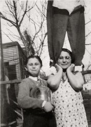Photo from 1927. This is my father, David Berger, at the age of 10. He came from a Brooklyn, N.Y., from a family that had a weakness for jokes and humor of every type imaginable. All of his siblings, with one exception, were natural comedians, offering the world laughs and smiles every chance they got.  The fact that his mother, my Granny Rose, agreed to be a part of this "unusual photo," combined with the mischievous look on Dad's face attests to that. (The owner of the legs standing atop Rose's shoulders is lost in anonymity). I have no idea exactly what Dad is holding, but I'm thinking it's an equally young Stanley Cup.
My very special and unique father died in 1973 at the age of 55. View full size.
ClotheslinedRe "The owner of the legs" ... Those pants are (obviously) empty. It must be laundry day, your dad is holding a Clorox jug.
Ahhh ... Clorox!Thanks, AT, for identifying the "Stanley Cup" for what it truly is. 
The leg thing was a joke of sorts. The first time I saw the picture, I genuinely thought it was a person standing on what appeared to be a none-too-wide support at the top of the fence. 
The realization that it wasn't someone at all came when I discovered the unbuckled pants. And thus, when displaying the picture thereafter, I would play it straight and bemoan the fact that I couldn't figure out who was "standing on Rose's shoulders."
But I was truly mystified about what Dad was holding. So, I now know more than before.
Thanks!
(ShorpyBlog, Member Gallery)