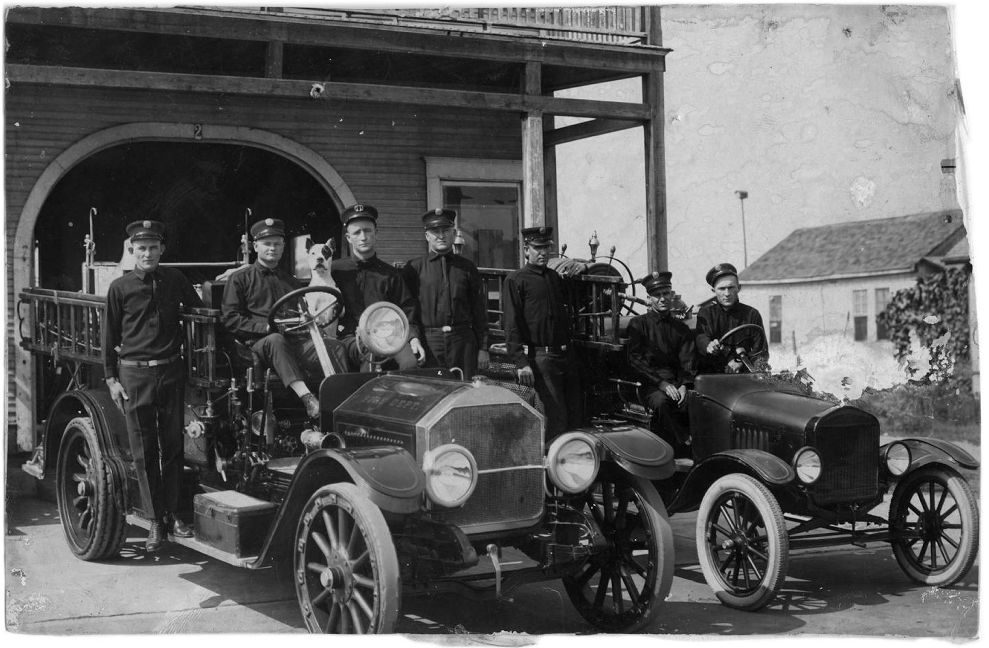 Photograph of seven men and one dog posing on fire trucks outside of the Number Two Fire Station on the corner of Thomas Boulevard and Houston Avenue in Port Arthur Texas. The trucks are a 1923 American LaFrance Pumper and a 1923 Model T Ford. The men are identified as Bill Woods, Ed Sykora, Eugene Ray, Shorty Robins, Jim Salyer, O'Neil Borel, and Pat Etheridge. View full size.
