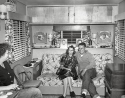 My good friend and former neighbor Jim and his wife Lois in their trailer home in the early 1950s, when he was stationed in Florida as a radioman in the Navy. He loved motorcycles, hot rods, music, cats, exotic birds, and most of all people. Talking to him was guaranteed to make you smile. Godspeed Jim! View full size.
From the pictureI will infer that Jim also loved boat building, nuts, family and - especially - Lois.
Redundant Lighting?I wonder what the plan was for the two lamps next to the wall sconces? Also, is that a waffle iron on the same shelf? It sure looks like the one we had, when I was a kid.
What a contrastto the previous photo ("Little Girl Blue"); these folks really look comfortable with their arms around each other, laughing, leaning IN towards each other, surrounded by family photos etc, and yes - smoking cigarettes.
TributeWhat a lovely tribute to your friend and neighbor. 
&quot;Silent Butler&quot;Note the "Silent Butler" on the right side of the ledge in back of the happy couple.  This contraption looked like a small covered frying pan with a wooden handle.    One simply dumped the contents of overflowing ash trays into the pan.  The ashes and cigarette/cigar butts were hence kept out of sight--and smell--until the lady of the house was ready to discard the mess into the garbage.  I have one of these relics packed away somewhere in our basement.
Waffle iron?Nope, I think it's what you empty ashtrays into before throwing the butts away?  Not to be indelicate, but it looks like a maternity outfit that she's wearing.  My mom had some that were very similar. Am I right? Thank you, Cazzorla, for sharing this peek into your friend's lives.  Everything about this trailer is perfect and I would have loved to have been invited over. Lois took pride in a tidy home, didn't she? Do you know if the trailer still exists?  It's a dream! 
TrailerKstan, I agree, it looks like it was a great trailer. They sold it decades ago.
(ShorpyBlog, Member Gallery)