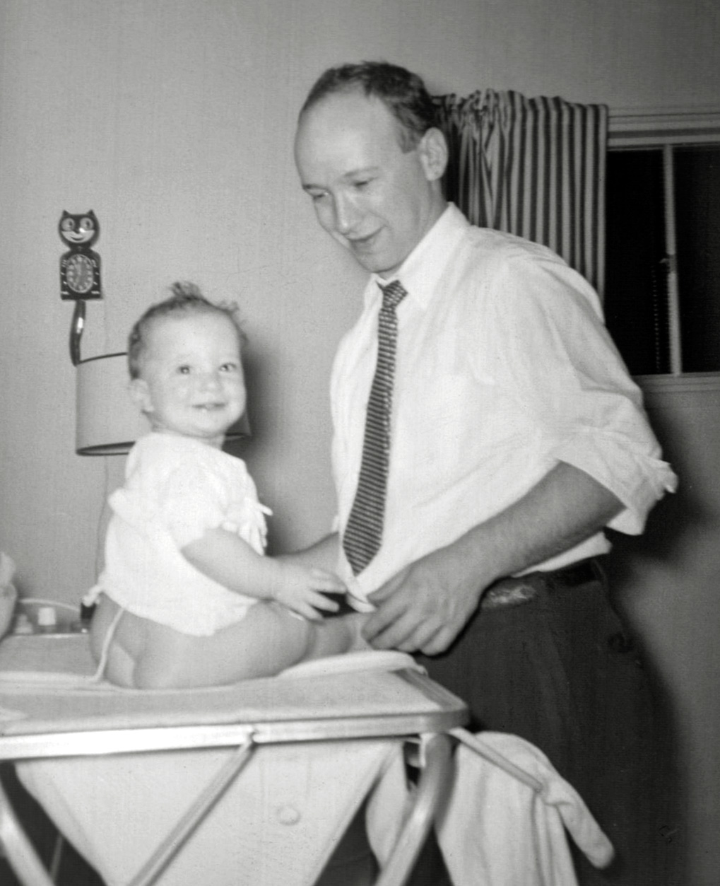 My mother snapped this picture of my father doing diaper duty with six -month-old  me. What I like about the picture is the paranoid schizophrenic Kit Kat Clock on the wall behind us. A rare bit of whimsy in my room, it clicked quite loudly as its slit eyes rolled left, then right, while its pendulum tail swung. Originally designed by Earl Arnault in 1932  for Allied Manufacturing Company, this original plug-in version was made from the thirties into the fifties. The earliest ones have only two lower paws. This one has four, indicating that it was from the end of the Allied era.

Kit Kat clocks enjoyed a pop culture revival in the 1980s and are still made today by the California Clock Company. Contemporary editions are battery operated, say Kit Kat on their face, and include a necklace or bow tie that none of the original clocks had. View full size.
