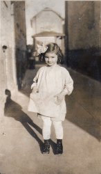 This photograph of my mother was taken in 1928 in Brooklyn, New York in an alley off of East 3rd Street. View full size. 
Oh, My!What a sweetie pie.  This little girl is the prettiest child I've ever seen.  
10/10That is a remarkably great photo.
The dramatic shadow, the dark alley outlining the sunlit subject, the short depth of field. Either a lucky snapshot or a carefully posed masterpiece.
On top of that, we have a cute model with her eye set straight on the viewer.
Super!
Lovely!An absolutely beautiful child. I love the fact that she's placed her little hands in some very little pockets.
Charming!I misread that as charming Sidelle Shear, and she is charming!
Beautiful Pic!She must've grown up to be a model or a movie star or something.
(ShorpyBlog, Member Gallery)