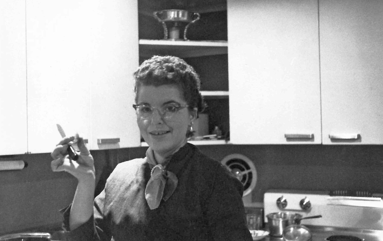 My mother poses with a small paring knife and a slice of apple in her hand (not a cigarette!) in the kitchen of our first home, which was in Levittown, Pennsylvania. The white steel cabinets, all-electric appliances and stainless steel counter tops are as mid-century-modern fifties as can be. So are her very fifties eyeglasses. That she is wearing a cravat at her neck, and jewelry, was not for some special occasion. She dressed that way every day.