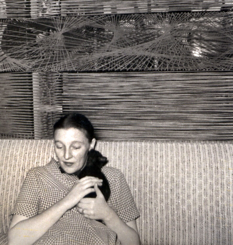 My mother, pregnant with me in Wakefield, Quebec. This was taken perhaps in July or August, 1956. I was born in mid-September. My father, who was an artist and had lived in Greenwich Village and the Lower East Side, moved my mother up from Ottawa to Wakefield where he planned to start an artist colony.
The studio was an old log cabin. This is in the house. The window covering is made from string and nails, like string-art but on a grand scale. The cat is either Henry or Midge. The night they brought me home from the hospital, Henry and Midge took up watch at head and foot of my crib. Mom is now 85. View full size.
