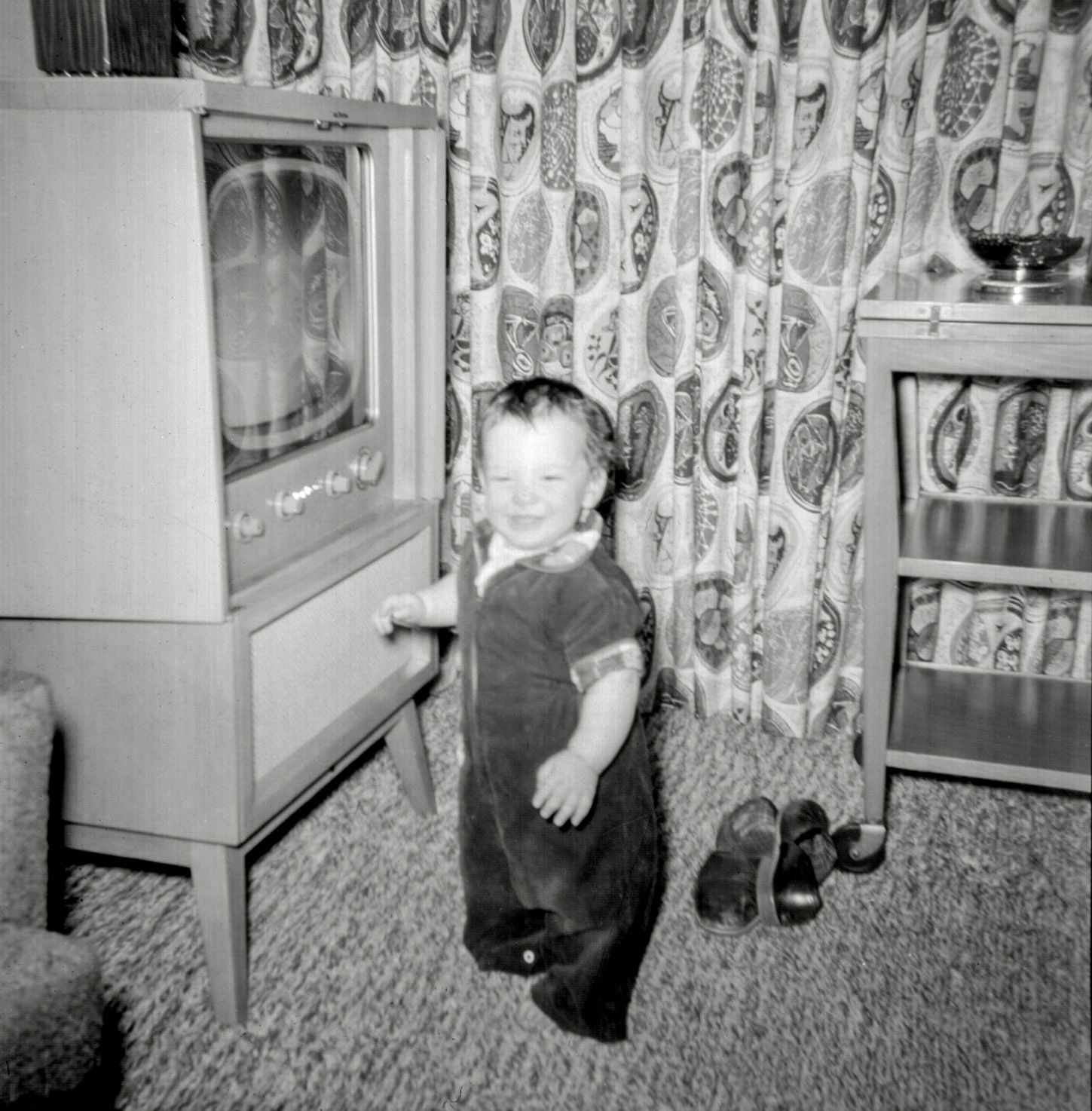In a previous picture I mentioned our family TV. But in that picture, the doors to it were closed, making it into a piece of blonde wood, mid-century modern furniture. It was a 1951 model RCA Kendall 17T174. We only had RCA TVs because my father worked at RCA Laboratories, in Princeton New Jersey, where he was involved in the development of color television. His slippers are on the floor behind me. To the right of them is a wheeled piece of blonde wood furniture that my mother called a tea wagon. Behind them all is a closer look at the very fifties fabric of the curtains my mother sewed for that living room. View full size.