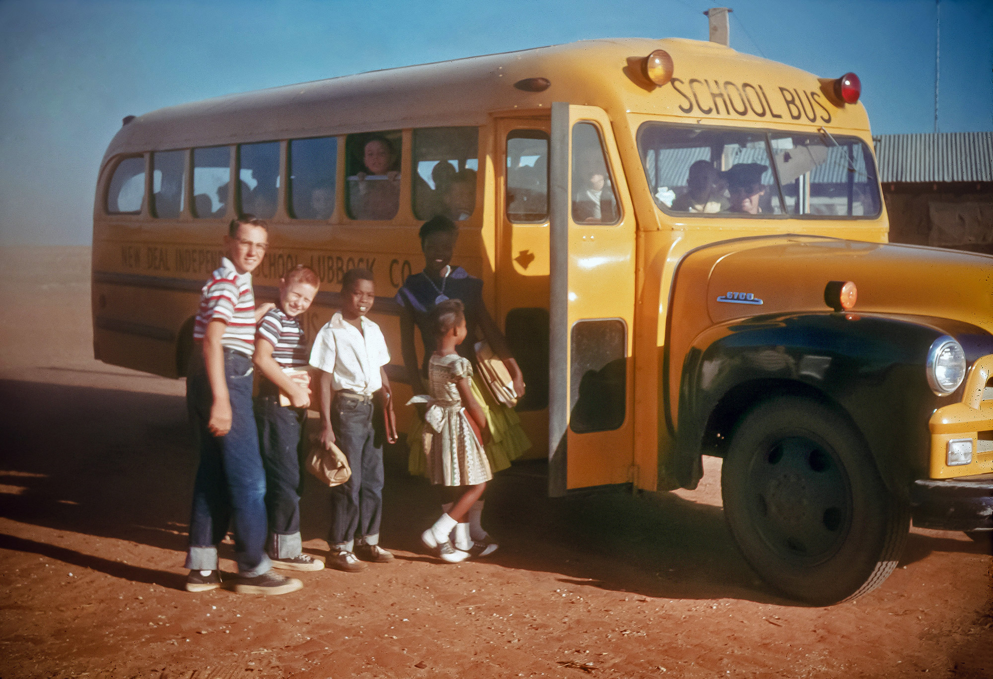 My dad (red-headed flat-top kid) with his books in hand, squinting from the sun, and his brother (taller white kid), about to board the school bus with their fellow classmates in New Deal, Texas in 1957. View full size.