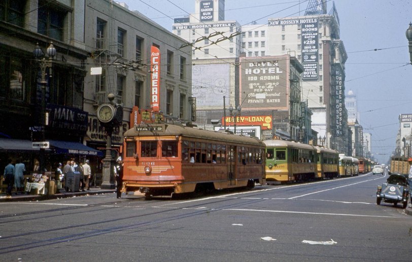This photo was taken by my close friend and fellow transit historian, Kenneth L. Douglas (1929-2017), who was born and raised in Huntington Park, Los Angeles. Ken gave me this 35mm slide about 1959, at the time I first became acquainted with him while we were both residents of Philadelphia.
The photo looks north at the intersection of Main and 6th Street opposite the main terminal of the Pacific Electric Ry.  The clock registers 2:30 PM and the slide is dated August 22, 1950.  A sharp eye will see LA City Hall in the distance at 1st and Main, and learn a lot about the state of the economy in the day.
Two different gauge tracks visible are used by two different trolley companies, Pacific Electric (the red cars) and Los Angeles Transit Lines (the yellow cars) along with overhead power to a trackless trolley (fourth vehicle in line), a diesel bus (fifth in line) and various other modes of transportation of the day.

