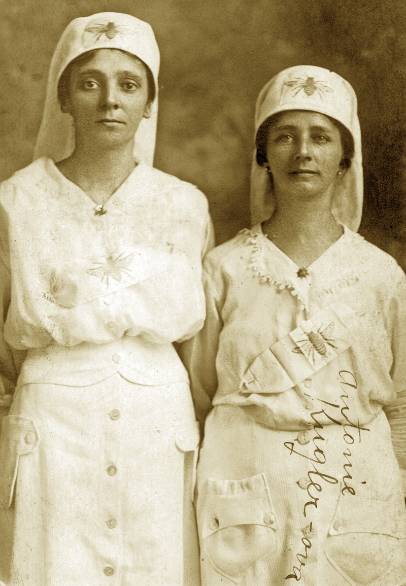 WWI-era photo of my paternal grandmother (R) and an unnamed colleague (L). Best I recall from verbal family history, the women were members of a volunteer unit preparing bandages for shipment to medical aid stations or hospitals in France.  Notice the bumble bee insignia on the caps and the sash.  If anyone can provide information on the volunteers or the insignia, I would appreciate it. View full size.

