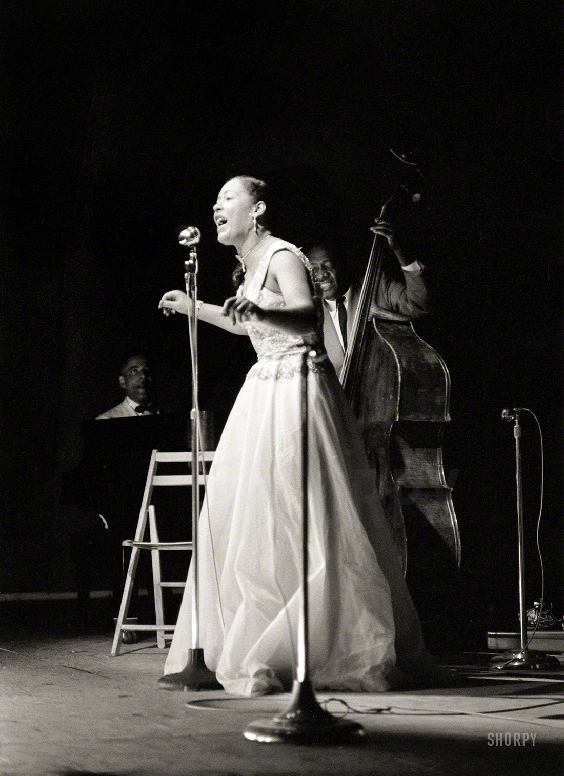 Billie Holiday, with Teddy Wilson at the piano and Milt Hinton on bass, at the Newport Jazz Festival in July 1954, five years from the close of her career and the end of her life. 35mm negative by John Vachon for Look magazine. (Caption updated thanks to our knowledgeable commenters.) View full size.