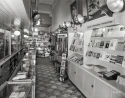 April 1, 1938. San Francisco. "Camera Craft store, 425 Bush Street. Mr. E.R. Young." 8x10 inch acetate negative by Moulin Studios. View full size.
Elementary PhotographyThoroughly Stimulating, Expertly Written.  For the Student, the Club Member, and the Amateur Photographer.  By C.B. Neblette, Frederick W. Brehm, and Everett L. Priest (Macmillan, 1936).  Under glass on the left, and in the middle bookshelf on the right, under the heading Latest Photographic Publications.
A professional and hobbyist dreamThis must be the most exciting camera store I have seen. So many periodicals. Looks like a hobby I would totally take up. Photography today means a smartphone app and a YouTube "how-to" video.  Not nearly as interesting at all.
Looks familiarThis looks pretty much identical to any of the many Embarcadero photo stores I recall seeing during my visits there.  But no doubt lacking the ridiculous markups and high-pressure salesmen of the more recent establishments.
Champlin On Fine GrainBy Harry Champlin, published by Camera Craft Publishing Company in San Francisco in 1937.  A September 1981 article in Popular Photography noted that Champlin “took a dim view” of the future of 35mm film since it was too small for “the exacting demands of commercial photography and newspaper work.”  He predicted 70mm as the new standard.
A Dearth of CamerasFor a camera store I certainly don't see many.
[Camera Craft was a publishing company. -Dave]
Brand NewThat looks like my Solar brand enlarger head behind the Popular Photography mag.
Camera stores!Isn't it odd that the photo hobby has changed so much? In my area, I would visit Penn Camera, Ritz Camera, and Industrial Photo every week. Didn't need anything. Just hanging out with my friends who worked there and messing around.
Days gone by. I used to love hanging around typewriter and watch repair shops, too. Chatting with folks with specialized, exact knowledge and geeking out. 
Those days were such fun.
Where&#039;s the &#039;Phone?It's at the rear of the store, attached to a set of shelves. It appears to be a Western Electric Model 211 Spacesaver, and in this store that really is a plus. 
When I was 12 my father gave me an Australian Kodak contact print set to make my own photos. It included a safe light, developing tank, trays, paper and chemicals to learn photo processing and printing. I later belonged to a camera club at school, and as an adult acquired an enlarger. I made my own prints in B&amp;W for many years, but never graduated to colour. The enlarger resides in my attic since I went digital. I can now scan my old B&amp;W negatives, and process them on my computer without all the liquids, fumes, etc. But I do miss the experience of visiting the camera store to stock up on supplies. 
Camera Craft Still being published.  Or rather being published again.
http://cameracraft.online/
Clamp lightsStill unchanged after almost 80 years!
Popular Photography 1937-2017Camera Craft may still be publishing but Popular Photography (bottom of the magazine rack) just shut down.
The Kodak LetterCurious about the "Kodak Letter" poster partly seen on the upper right, I found it was a 1918 magazine ad encouraging people to send photos to their soldiers "over there" in WWI. So it was already a vintage poster (and camera) in 1938.
[Good work. I was wondering about that thing that looks like an antique cable modem! - Dave]
Camera in &quot;The Kodak Letter&quot; adThe camera in the "Kodak Letter" poster from 1918 probably is
this one. It does look a bit like a cable modem when folded! I recall my granddad (who was just a bit too young for WW I) had something like this when I was small. It was fascinating.
[However, many Kodak folding cameras of the period looked like this when closed. -tterrace]
Interesting location.The location of this business would later become the location of KFRC, the legendary San Francisco radio station of the 60's and 70's.
(The Gallery, San Francisco, Stores & Markets)