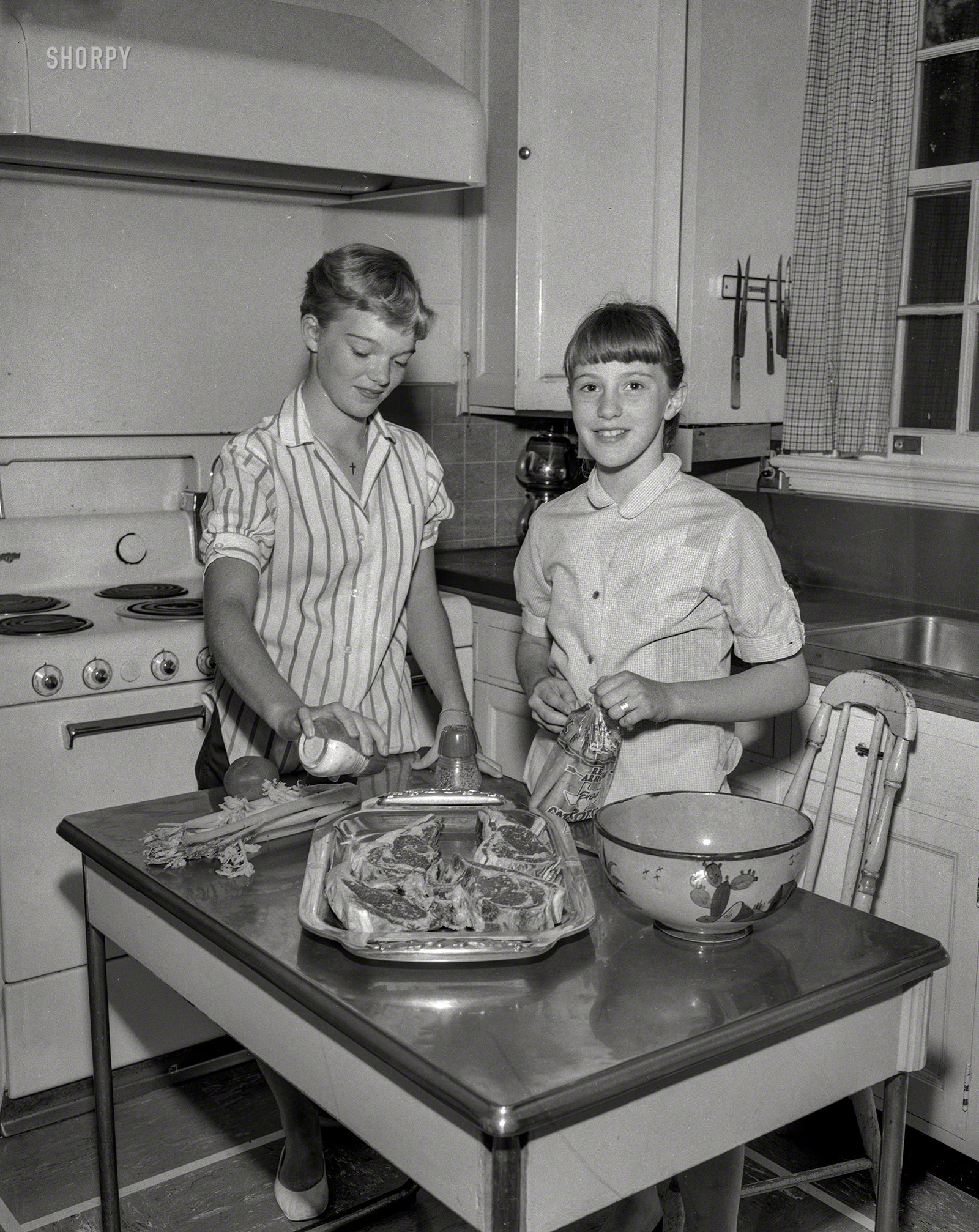 From circa 1956 and Anytown, USA, comes this captionless snap of two young ladies and a broiler tray of chops, next to a chair with our name on it. "Bone" appetit! 4x5 negative from the Shorpy News Photo Archive. View full size.