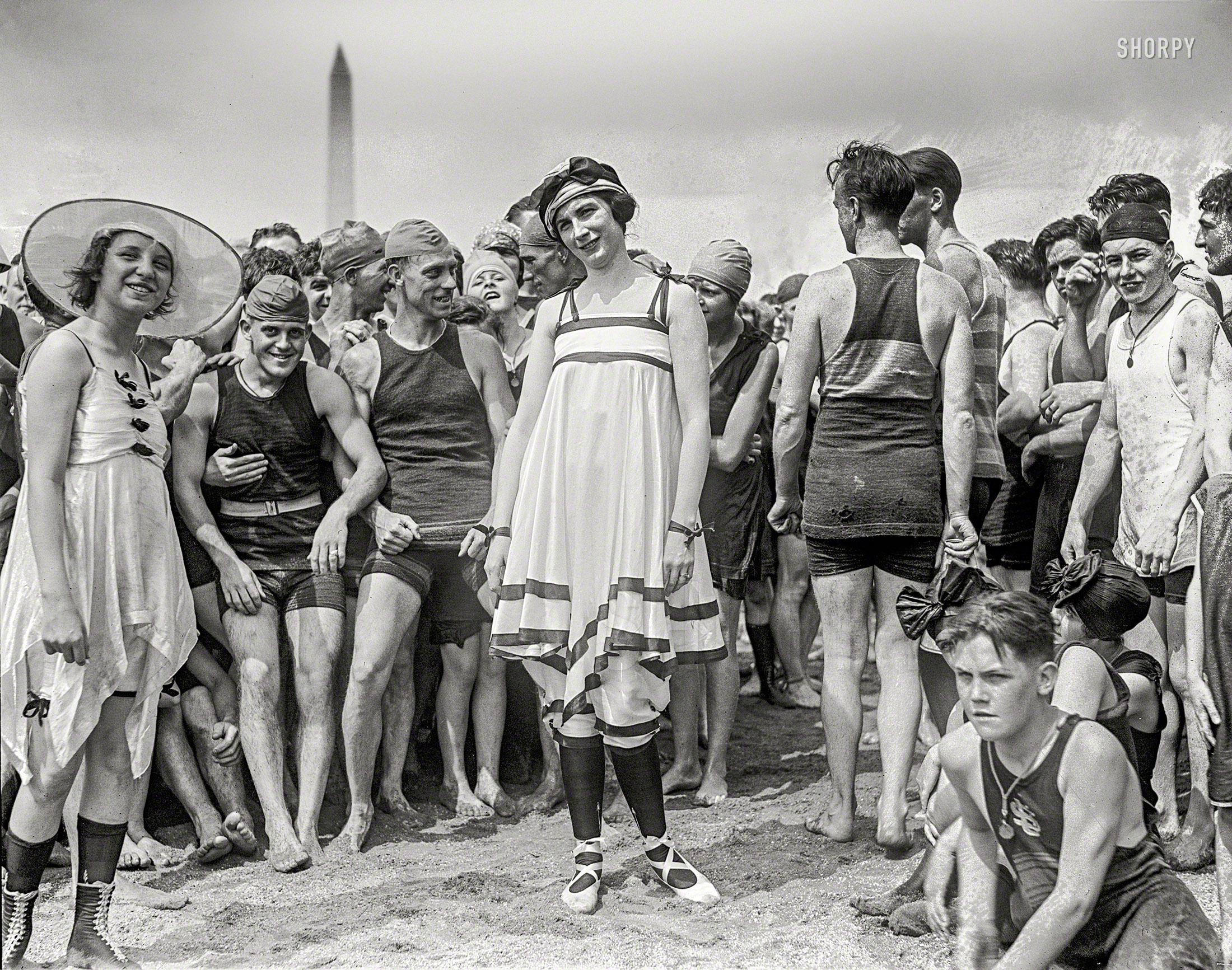 July 26, 1919. Washington, D.C. "Bathing beach parade at Tidal Basin." National Photo Company Collection glass negative. View full size.

GALAXY OF BEAUTY PARADES AT BEACH

Comely Damsels in Scant Attire Win Prizes for Their Appearance.

&nbsp; &nbsp; &nbsp; &nbsp; JULY 27 -- While more than 5,000 persons clambered to each other's shoulders and to roofs of nearby buildings to view the Annette Kellermanns at the first annual beach parade at the Tidal Basin yesterday afternoon, Mrs. Audrey O'Connor, 620 Maryland avenue southwest, was proclaimed by the judges as Washington's most beautiful girl in a bathing suit. Mrs. O'Connor wore a blue and orange jumper, blue cap and orange tights. Miss Dot Buckley, 1250 Tenth street northwest, received honorable mention in the contest. Her suit was a creation in red, white and blue.
&nbsp; &nbsp; &nbsp; &nbsp; First prize in the costume contest was awarded Mrs. Grace Fleishman, 5 Iowa circle, who wore a white silk suit with black and white border and a white silk hat. Miss Muriel Gibbs, costumed as Miss Liberty in stars and stripes, received honorable mention. Silver loving cups were awarded to the winners of both the beauty and the costume contests. 
&nbsp; &nbsp; &nbsp; &nbsp; Following the parade of the score or more of beauties between cheering crowds of bathing beach fans, the former faced half a dozen movie machines and a battery of press cameras. Later one of the winners obligingly did a modified "shimmy dance" for the movie men.
