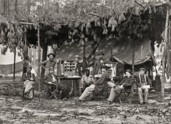 August 1864. "Camp of Chief Ambulance Officer, 9th Army Corps, in front of Petersburg, Virginia." Albumen print, photographer unknown. Civil War Glass Negatives and Prints collection, Library of Congress. View full size.
Photoshop not around in 1864The fellow standing at the left leaning on the cabinet seems to be missing his lower torso and legs, and has a tree where they should be. 
[That's his stump. - Dave]
Pine pergolaThat's a clever structure they've put together of pine logs and boughs. I'll bet it smelled sweet and made a swishy sound in the hot summer breeze. In other news, the young man seated on the far right looks like he was about fourteen years old. I hope he made it home after the war.
Function of an ambulance campBoy, that's an interesting photo.  Is there any identification of the men
in the picture?  Does the "ambulance officer" have to do with evacuation
of the wounded, as it would seem from today's idea of an ambulance?
The camp appears to include a sorting station for mail and messages.
Post Office?Fascinating photo. I know the caption says Ambulance Officer, but this looks more like the camp post office. Two of the standing men are holding almost identical packages. Maybe some Civil War buff can guess what would be in them.
[The caption says this is *the camp* of the Chief Ambulance Officer. The larger camps having their own telegraph offices, messengers, letter carriers, etc. - Dave]
Legs like a treeIf he was missing both legs, why would he still be in the military? Why wasn't he sent home? Or to a soldiers home? This picture is going to disturb me all day.
[He's standing behind the stump. - Dave]
Battle of the CraterThis must have been taken shortly after the fiasco of the Battle of the Crater, but maybe also after the Second Battle of the Weldon Railroad, which was more successful. I can't even imagine what these men had just seen.
Multiple usage of equipmentLooks like the bone saw served double duty as the pine limb trimmer for the camp. 
Civil War CelebThat’s got to be a relative of Ed Norton, second from right. In a director’s chair no doubt!
Eyes have itIntense imagery. Everyone, even the kid on the right, has the same look in their eyes. The black man is the only one with anything resembling a smile.
What are they hiding from? The infamous Confederate Air Force? Or at least the Viginia Balloon Corps? 
Unhappy lotGrumpy bunch of guys, but Dave's response to the post by FixIt gave me a belly laugh to make up for the gloom!
Fancy folding chair Seated second from the left.
(The Gallery, Civil War, Medicine)
