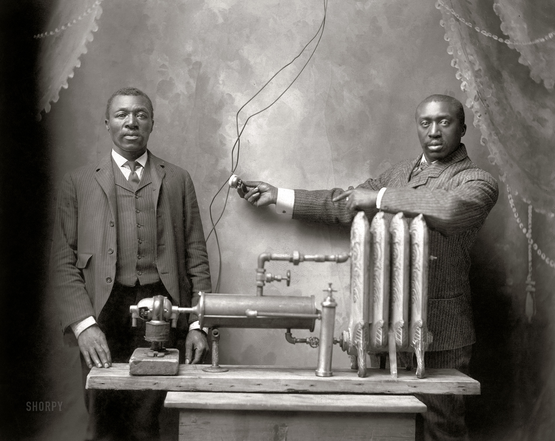 "Start of super heating union. [Inventor Charles S.L. Baker and another man, possibly his brother Peter, standing behind heating (radiator) system.]" Photo by Bode, Fifth and Felix Streets, St. Joseph, Missouri. Copyright by Charles S.L. Baker, Feb. 12, 1906. View full size.