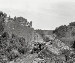 Circa 1899. "Near Lewiston, Minnesota -- The Pulpit." Yet another rock formation with a fanciful name. 8x10 inch dry plate glass negative. View full size.
Link and Pin CouplerThat locomotive is equipped with a primitive Link and Pin coupler. You can see a link hanging from the pin, plain as can be.  This is a dangerous manual coupling system that exposed RR workers to dismemberment and death. 
There is no air brake hose visible, either. 
The 1893 railroad safety appliance act made automatic couplers and air brakes mandatory. It was phased in over seven years, so the actual date of the photo is most likely before 1899 and certainly is not after 1900.
Karst TopographyI'd like to have a look in that cave on the right.
Does anyone know if the Pulpit still exists?
Filling in a trestle.It was common practice when these rail lines were built to erect trestles over low places and when the line was completed and in service go back and fill in around the trestle. This is a good picture of that being done. 
Special Train?The engine is a Chicago &amp; North Western S-2 class 4-6-0 built in 1890.  As such it was an example of the biggest and best the railroad had before 1900. The engine lasted until 1925 when it was scrapped.  In this photo it hauls one passenger car--an attendant appears looking out from behind the car suggesting a very  special train. Perhaps top management. Perhaps an inspection train. The locomotive is a good deal more powerful than needed for one passenger car--an ornate at that. 
Special Train followup. Responding to SouthBendModel34--The Chicago &amp; North Western (C&amp;NW)line through Lewiston I believe started life as the Winona &amp; St. Peter Railroad which had a close association with the C&amp;NW.  Note the C&amp;NW logo  on the front of the smoke box of number 148. Construction was started about 1866 and the C&amp;NW purchased the railroad in about 1867 though it operated it as a separate entity until 1900. The railroads shared motive power designs and classification codes though motive power , numbering and logos on locomotives were kept separate until the final merger.    
Locomotive 118The number plate at the center of the locomotive smokebox seems to read 118.
The line through Lewiston MN is currently the Dakota, Minnesota &amp; Eastern, which is a successor to the Chicago and Northwestern Railway.  (Which may or may not have constructed this line: can any Shorpy Sleuth come up with a construction date for the RR through Lewiston MN? 
The trestle fill-in would probably be not too long after the opening of the line.
Can photo enhancement read a RR name on the side of the tender, or the name of the single passenger car? (Which is probably the RR's Business Car. This looks like an inspection trip to check on the progress of the trestle fill.)
There's curved, raised lettering on the locomotive smokebox which I would guess to be the name of the locomotive. builder. Can that be enhanced ?
[Signs on the tender and car aren't legible. -tterrace]
PulpitThe cave on the right is a legend about it being the Devil's Hole. (see photo)
[The photo you attached related to the Lewiston in New York state, not Minnesota. -tterrace]
[Thanks. Being a Brit, I don't know my NY from my MN...]
C&amp;NW Business car 104The open platform car was leased (owned?) by the Detroit Photographic Company.
(The Gallery, DPC, Railroads)