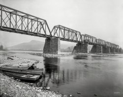 Circa 1901. "Bridge over the Susquehanna at Pittston, Pennsylvania." 8x10 inch dry plate glass negative, Detroit Publishing Company. View full size.
The view today courtesy of GoogleView Larger Map
Why is the bridge on one side of the supports?I am wondering why the bridge is on only one side of the supports. Would another track have been put beside it eventually? Or was this how they were constructed-- I notice that the side holding up the track seem reinforced with stone coming down at an angle.
Fishing For?Bullhead catfish or sunfish I'm guessing, since he's still-fishing.  Bullheads on bottom, sunnies if he's using a float.  The smallmouth bass so sought after on the Susquehanna these days had probably not been imported yet--they're not native.  Carp were only beginning to become unwanted guests in America's rivers.
CamelbackThe locomotive on the bridge is a type called a Camelback.  The engineer and brakeman sat in a cab that straddled the boiler in the middle of the locomotive.  The fireman had his own shelter at the rear to transfer coal from the tender to the boiler firebox.
The reason for this design was that they burned local Anthracite coal which is very high in fixed carbon.  It burns hot but slowly, like charcoal.  To get enough heat to power the locomotive the firebox had to be almost as wide as the locomotive, making it impossible for the engineer to see ahead from a rear mounted cab.  The solution was to put the cab ahead of the firebox.
I&#039;ve fished exactly where he is, probably 80 years later.Technically this fellow is sitting in West Pittston, not Pittston itself, which is what you're looking at across the river.  West Pittston and Pittston are two separate towns, with different school districts.  So we Wyoming Area Warriors considered the Pittston Patriots our sworn enemies.  Of course back then, the worst you did to your enemy was maybe TP the trees in their front yard.  That always showed 'em!
Regarding the fishing, he's probably fishing for his dinner. Many years later, we caught a lot, but never ate them, since the river these days is pretty, but not necessarily something you want to eat out of.
And one final thing for jaylgordon-the bridge abutments are slanted like that on the upriver side, to allow debris flowing downstream to more easily roll off and not get stuck on them.
Bridge SupportsThe Pennsylvania Railroad and her subsidiaries would typically build bridges in this manner, with a piling wide enough to accommodate two tracks; but only place one track over the bridge.  The reasoning, at least for the railroad, was that if traffic or demand ever became such that a second track needed added; it was simply easier to plunk a new bridge down on the extra width. This also meant that the existing line would not need to be closed during the construction.
The locomotive in question looks to be one of the PRR's 4-4-0 camelback types, though it's possibly a 4-6-0. (Can not tell from the angle.) Here is an image of the possible locomotive type.
(The Gallery, Boats & Bridges, DPC, Railroads)