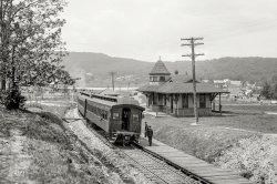 Washington County, Maryland, circa 1905. "Buena Vista Springs station at Pen-Mar." A Blue Ridge mountain resort, developed by the Western Maryland Railway, that took its name from the two neighboring states. 8x10 inch dry plate glass negative, Detroit Publishing Company. View full size.
Handbrake on Passenger Car. Handbrakes are still used as 'parking brakes' and occasionally to add drag to remove slack on freight cars when switching. 
Backup brakingThe passenger car has both a hand operated brake wheel and an air brake hose.  
Either it's an older car that was refitted with air brakes after going into service, or railroad management was not quite ready to place full faith in Mr. Westinghouse's new-fangled system.
Backups are StandardHand brakes are still installed on railroad cars. It is not because of not placing full faith in the air brakes. When disconnected from an air source, the air will eventually (hours to days) leak away, rendering the air brakes useless. 
Each car has an air reservoir (actually, two reservoirs), which is filled from the train air line (the smaller hose in the picture). As long as the pressure in the train line is maintained, the brakes will not apply. When the train line pressure is reduced, a (fairly complex) valve will allow air from the reservoir to enter the brake cylinder, applying the brakes. Up to a point, the amount of air admitted to the brake cylinder is in proportion to the pressure reduction. If the pressure reduction is large (to zero, for example) or quick (due to a broken hose, for example), all of the air from both reservoirs is admitted to the brake cylinder, resulting in an emergency brake application.
But, the air can leak away, so if a car is to be left standing, its hand brake needs to be applied.
Hand brakeAll railroad cars, even the newest, have hand brakes.
These are required to apply or release the brakes on a car or string of cars, not connected to the engine or other source of air.
Lots of RR DetailsOn the left side of the train is an early signal with a black "target" with a lighter-colored center, probably white.  The arm pivots up or down to indicate "proceed" or "stop".  The up position is usually the "proceed" aspect - derived from the previous "ball signals"
In front of that is a "harp" style switch stand for moving a track switch.  (Note that there are two tracks up ahead of the train, which is standing on a single track.)  The target on the harp switch lever is round with a cross on it.
Beyond the signal is an edge view of a road crossing warning sign. In those days, the crossbuck style was not yet universal.
At the rear of the train, there are two hose couplings, one for the air brake and one for steam heat. (The ventilators passing through the roofs of the coaches are for the lavatories.) 
The peak of a smaller building is visible to the right of the train. This might be a signal cabin.
The depot itself has absolutely delicious architectural detail in a "shingle style".  Note the eyebrow window in the roof, the octagonal tower, and the corbelled chimney.
The bay window muntins are certainly very ornate; a pattern of small squares surrounds the larger panes.
The depth of field of these large-plate cameras never ceases to amaze, does it? 
And the House on the Left....is still there.

CouplersThe is another interesting detail clearly visible in this remarkable photo.  The year 1905 was within a transition period of several years where railroads handled a mixture of passenger and freight cars that were slowly and gradually becoming equipped with the much safer Janney or modern knuckle type couplers.  These replaced the old fashioned and very dangerous link &amp; pin type coupling method that was responsible for so many railroad trainmens loss of hands and/or fingers.  Clearly visible in this photo is a small horizontal slot in the closed knuckle at the end of this passenger car.  When necessary, any railroad car that still retained the old link &amp; pin equipment could be coupled to this passenger car by the old system of the trainman guiding the link into that slot, and dropping the pin down through the hollow knuckle and through the link.  It took quite a few years before all of the old link &amp; pin equipment was gone and the new Janney coupler knuckles no longer had to be provided with the slot and the vertical hole and old fashioned pin.   
Mail catcher at restThe white device to the left of the train is what is known as a mail crane. The postal worker would climb the steps and mount a bag vertically so the hook arm on a Railway Post Office car (or portion of car) could capture the bag as a train passed without stopping. 
In front of the mail crane is a "harp" style switch stand. These were associated with the "stub" style turnout that preceded the tapered movable points we know today. The stub switch had the main track rails move to align with whichever route as chosen.  
Station is long gone, but. --the flat roof building in the rear is still there and is now the American Legion hall. It is somewhat expanded but retains the roof line and the shed extension in the rear. The station would have been to the left of the crossing sraight ahead.

(The Gallery, DPC, Railroads)