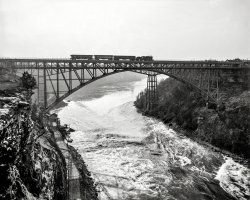 Circa 1900. "Niagara Falls, New York. Whirlpool Rapids (Grand Trunk Railway) Bridge with Michigan Central Cantilever Bridge in background." 8x10 inch dry plate glass negative, Detroit Publishing Company. View full size.
Conductor to apprehensive passenger:"If we derail, don't worry about drowning; the fall will kill you."
Awesome!Awesome photo of awesome features. Wow!
Bridges &amp; TrainsLooking southwards/upstream, with the USA on the left and Canada on the right. The falls are about 3 kilometres upstream from here.
The tracks on the left at the riverside are part of the Niagara Gorge Railroad, an electric line that ran from 1895 to 1935.
The bridge may be owned by the Grand Trunk, but that's not a Grand Trunk train. The lead locomotive is a camelback, and the GTR didn't have any. This may be a Lehigh Valley locomotive, as there was transfer traffic between the LV and the GTR over the bridge
Still in useThe bridge continues to carry both trains and on the lower deck, auto traffic; but no longer pedestrians.
It's been designated a special NEXUS pass bridge here in Niagara Falls. Shame because walking across the bridge as a boy and teenager, it was thrilling to see the Niagara River Rapids swirling below through the spaces of the wooden planks on the pedestrian walkway.
A Century-Plus LaterThis is a view from the Canadian side from October 2011. The bridge behind the Whirlpool Rapids Bridge is now the Michigan Central Railway Bridge which replaced the Michigan Central Cantilever Bridge in 1925 (and has itself fallen into disuse.)
Another railroad below itWhat is the railroad at river level? and what it the infrastructure above and next to it?
(The Gallery, Boats & Bridges, DPC, Railroads)