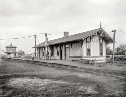 Circa 1904. "Depot at Braidwood, Illinois." 8x10 inch dry plate glass negative, Detroit Publishing Company. View full size.
Another name for BeeGuy&#039;s listAs a preteen I rode the GM&amp;O's "Gulf Coast Rebel" from St. Louis, MO to Waynesboro, MS a few summers without parental accompaniment.  (No detour to Braidwood.)
Still waitingFor the last train to Braidwood:

Now home to the Braidwood Historical Society.
Chicago &amp; Alton RouteOn the main line 57.3 miles south of Chicago. Also a junction with a spur off of a branch that ran between Joliet and Coal City.  Later this road became the Gulf, Mobile &amp; Ohio, route of such passenger trains as "The Abraham Lincoln" and "Midnight Special". 
DepartureThe depot was moved from its original location (the website doesn't say when although the photos of it being moved were uploaded in 2012)
4 Photos
Attractive SpiresThis prosaic building had been given a lot of interesting architectural details, such as the roof brackets, the bargeboards, and most of all the wooden spires.
Note that the two-spouted wooden water tank in the background has a matching spire.  Pretty spiffy ! 
The tall windows let in plenty of money-saving natural light.
The size of the building, the train order signal, the large doors, signage, and the two baggage carts, one standing ready at each end of the platform, suggest that this was a multipurpose building which handled train order operations, passengers, checked baggage, Western Union telegraphy, Railway Express parcels, and perhaps also Less-than-carload freight. 
It would be interesting to know more about the track served by the far water spout of the water tank. Was this just a siding, or was Braidwood a junction point? 
LCL freighthouseThe right half of the building was the “freight house.” The large door giving access provided ample room for the freight handlers (with a union craft of their own in later days) to wrestle large pieces of LCL freight (perhaps a piano from the Sears Catalog) into the structure until the consignee could arrange a pick-up. There is probably a door on the back side of the building that an LCL boxcar was spotted at for unloading larger pieces of freight. Large and cumbersome LCL freight required too much time to unload while sitting on the main, so the entire car was simply left to be unloaded at convenience. In railroad parlance, the track would have been called a “house track.” It would have a main line switch at both ends, making it possible for both northward and southward trains to spot and pick up as necessary. The water spout over it is a mystery.
Actually Braidwood was not a junction with any branch or carrier. The south switch for the branch to Coal City was at Gardner, and the north switch going back to the main was at Elwood. Braidwood was almost exactly half-way between those two points.  
(The Gallery, DPC, Railroads)