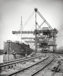 Circa 1910. "Brown electric hoist unloading freighter Constitution at Cleveland." 8x10 inch dry plate glass negative, Detroit Publishing Company. View full size.
That hopper carbelongs to the Pittsburg &amp; Lake Erie Railroad, seems they may be unloading (loading) coal.
The Constitutionwas a 379-foot long barge, launched 22 April 1897 at West Superior, Wisconsin, by the American Steel Barge Company, a firm better known for the design and construction of the unique Great Lakes "whaleback." The steamer Victorious generally towed the Constitution, both owned by Cleveland's Pickands, Mather &amp; Co. Brought back to Superior to be lengthened over seventy feet in 1905, Pickands, Mather sold the vessel to Cleveland's Pringle Barge Line in 1922, which had it converted to a self-unloading barge five years later using a unique system developed by noted Chicago marine contractor Jacob Sensibar.  The Constitution ended its career primarily hauling coal from Toledo to Detroit, towed by the big Diesel tug S. M. Dean, and was dismantled at Port Colborne, Ontario, in 1968.
Brown and out.The cables visible next to the tracks are probably there to move a "larry", sometimes called a "pig", a small rail car that connected to the hoppers, so that they might be moved on the wharf without the need of a locomotive.
NYPANO dacksThis looks like the New York, Pennsylvania &amp; Ohio dock on Whiskey Island. This facility, but not the pictured unloaders, lasted until Conrail was formed and then were surplus due to the huge PRR (Cleveland &amp; Pittsburgh) operation with its four massive Hullett unloaders just across the old river bed.
 Those Pittsburgh &amp; Lake Erie hoppers likely brought coking coal to Cleveland, and will get a load of ore for a return to the Ohio Valley- probably near Youngstown. 
(The Gallery, Boats & Bridges, Cleveland, DPC, Railroads)