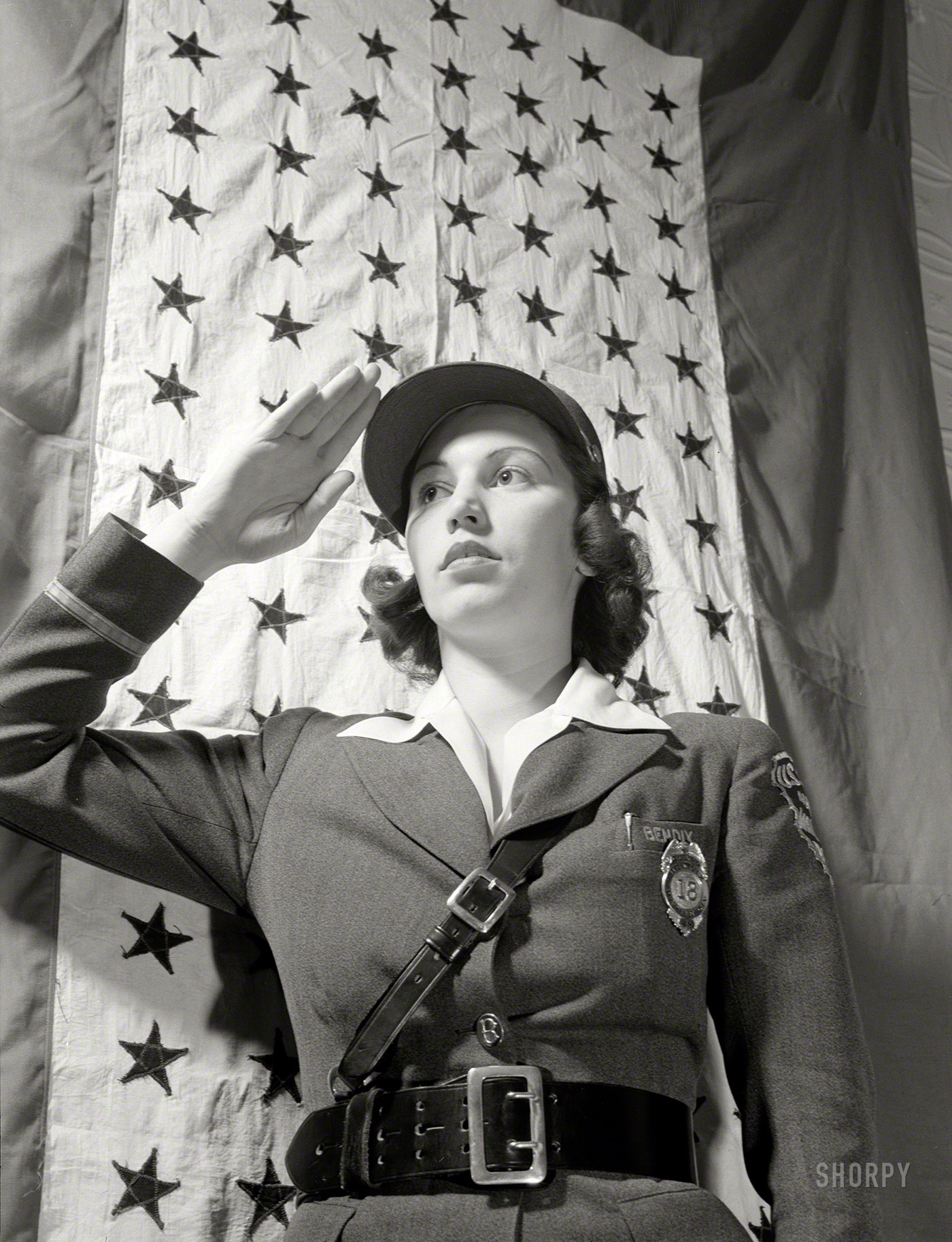 March 1943. "Safe clothes for women war workers. Estelle Hendel, twenty-eight, a guard at the Bendix Aviation Plant in Brooklyn, New York, stands before the company service flag and gives the correct salute." 4x5 inch nitrate negative by Ann Rosener for the Office of War Information. View full size.