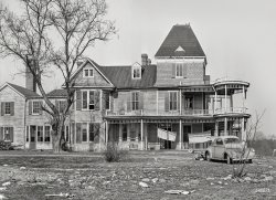 March 1941. "House being converted into a 'nightclub' near Laurel, Maryland." Medium format negative by Martha McMillan Roberts. View full size.
I see youEven the car looks sad. 
I wonder who the fellow is who is almost hiding behind the tree.
RegardlessThe corner porch detail is really a fun element.  
I&#039;m scared... of the scare quotes around "nightclub," though I agree with switzarch that the corner porch is is interesting.  It looks like a big place that could accommodate many "night activities."
Shorpy Vehicle Identification ImperativeThis one's easy: Willys Americar coupe.  The harder part: is it a '37 or a '38?  Perhaps a Willys expert could chime in with a ruling.
A swinging countyHaving spent about six months in Ann Arundel county in 1950, I vividly recall that cafes often had slot machines installed -- not the kind that dispensed coupons for free cigarettes, either.  I don't recall seeing them in Baltimore, but the officers' club at Fort Meade had them too.
Oddly, my mother would often give me a nickel to play the juke box, but not to play the slots!
More of a NightMareStraight outta Stephen King.  Bet there's a Pet Sematary 'round back.
Willys!I would say that car is a 1938 Willys Model 38 Coupe. Wish I had it in my backyard!
Dead CenterI like the three bullet holes in the ash can. Ready for Saturday night.
Gives Me the WillysI'll bet that the owner of that poor Willys wished, around 1944, that he'd taken better care of it. Amazingly battered up for a fairly late-model car. The name "Willys", contrary to popular belief, is pronounced "Willis"
Willys IndicatorsThe lack of a pronounced rain gutter above the door indicates that it is a 1937.  The two vertical front bumper guards (ignoring the large aftermarket one in the middle) indicates that it was of mid-to-late 1937 manufacture, as the early production versions (starting in the fall of 1936) did not have them.  The lack of three chrome trim spears on the sides of the headlight pods indicates that it is a Standard, rather than a DeLuxe coupe.  It sold for $499 new.
1939 WillysIn 1945 my parents returned from their World War II military duties in Europe, and my father was accepted into the Harvard Business School. Here they are ready to leave his mother's home on Cornish Road in Toronto for Wollaston, near Boston. The 1939 model Willys is not in bad shape compared to the one in the photo, and there has been a slight redesign of the body. Since it is a coupe, the trunk was jammed to overflowing, which caused a scene at the U.S. border. My father had no recollection of the hood "ornament". Upon returning to Toronto in 1947, when I was born, it was replaced with a 1929 Nash, but that is another story.
Beat up WillysThe Willys coupe is a 1937 or '38. If the picture was taken in 1941 that coupe has had a rough life. The coupe in Angus J's photo is a 1940, identified by the rectangular grille and chrome strips on the nose of the hood. The 1939 had  a grille similar to the '37-'38 but more pointed and the head lamps had a matching pointed glass.
Angus J&#039;s hood ornament. That's Ferdinand the bull.
 Normally depicted laying in or holding flowers in his mouth.
high wireWhat is the nature and purpose of the line stretched between the peak of the dormer and the cupola? Lightning protection? Some kind of radio antenna?
(The Gallery, Cars, Trucks, Buses, Martha Roberts)