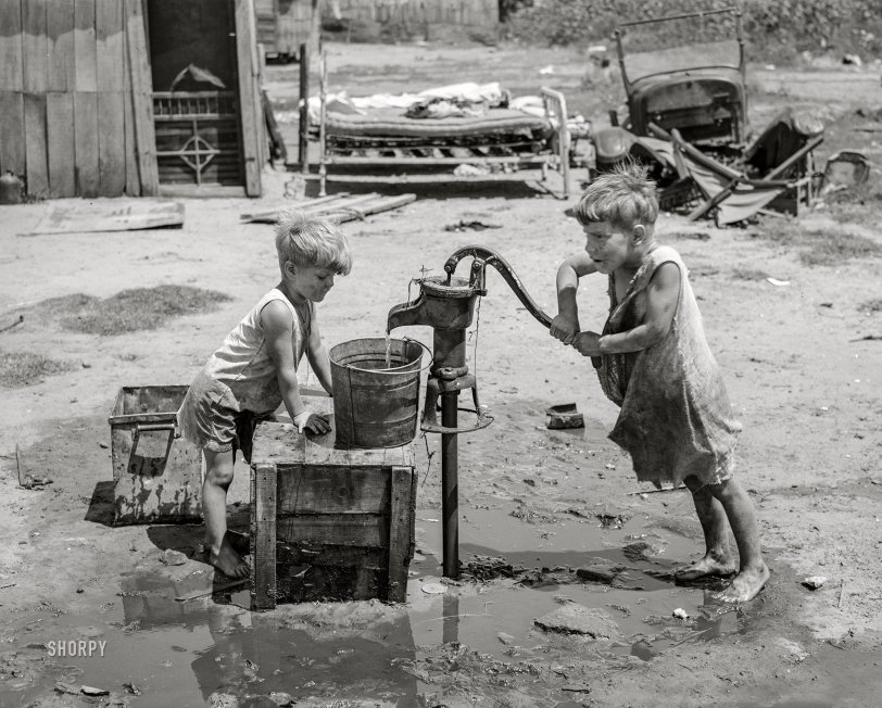 July 1939. "Children of May Avenue camp pumping water from thirty-foot well which supplies about a dozen families. Oklahoma City, Oklahoma." Photo by Russell Lee. View full size.
&nbsp; &nbsp; &nbsp; &nbsp; General caption: Photographs show exterior and interior of shacks, tents, other makeshift shelter in May Avenue camp, which is partially under bridge and adjacent to city dump and hog wallow. Squalor, filth, vermin in which poverty-stricken inhabitants dwell. Water supplied by shallow wells and water peddler. Piles of rubbish and debris in which children and adults have injured feet. Privies. Families eating food from vegetable dumps, packing houses and discarded from hospital. Children clothed in gunny sacks. Malnourished babies. Sick people. Cooking, washing, ironing, patching. Improvised chicken coop. Corn patch."
