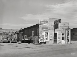 September 1939. "Market and grocery in Questa, New Mexico." Medium format negative by Russell Lee for the Farm Security Administration. View full size.
SignageI shutter to think what a picker wold pay for all those signs.
[It's "shudder." - Dave]
Equal relief for men &amp; ladiesIf the meat or vegetables are not all that fresh and end up causing digestive problems, thankfully there are two outhouses to run to at the back of the grocery shop.
Fully equippedFully equipped with all the modern conveniences... including facilities for Men and Ladies out back.
I wonder why they were called &#039;Cash Markets&#039;?In the town where I grew up there was a small store a couple of blocks from us, called "Scotties' Cash Market". It was was pretty small, but it had a lot of stuff packed in that space. Did a good business in the 1960s, but died a slow death in the 1970s when two things happened: (a) the town turned the street it was on (the Main Street) from two lanes with parking into four lanes and banned parking, and (b) a 7-11 clone opened down the street with off-street parking.
[It means you can't buy on credit or run up a tab. - Dave]
AvalonYou’d never guess they cost you less.
Everything a road traveler could need or wantOne very well stocked roadside market, cigarettes, tobacco, rolling papers, bags of flour, Coca Cola, free air and comfortable rest rooms. 
Those OuthousesI hope the meat coolers were on the other side of the store. 
Shorpy Parade of Pickups PastAbout a 1935 Dodle.  If you go much earlier in the 30s, the Dodles I can find pictures of had rear-hinged "suicide" doors, at least on the driver's side.  Of course, it's possible that Fiat Mercedes-Benz Chrysler Dodge Brothers built a pickup with a suicide driver's door and a regular passenger door, to clear the spare tire -- that would be a very Chrysler thing to do.
(The Gallery, Russell Lee, Small Towns, Stores & Markets)