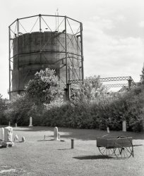 July 1938. "Gashouse and graveyard. Dover, Delaware." Medium format negative by John Vachon for the Farm Security Administration. View full size.
Raising the PressureThis photo is the first that I've seen that shows how (concrete?) blocks have been added to the perimeter of the top course in order to raise the gas pressure. These gasometers, or gas holders, acted like an inverted tin can in a bowl of water. More weight on top would cause an increase of pressure of the gas inside. As the gas was consumed, the inverted "can" would ride the guide rails and slowly descend.
(The Gallery, John Vachon)