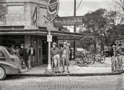 December 1940. "Soldiers on street corner in Starke, Florida. Boomtown in defense district." Medium format acetate negative by Marion Post Wolcott. View full size.
Piss off, Indeed (Plus added information)Dave,
That guy is wacko. I'm on your side.
My mother-in-law was a St. John and I thought this might be family. Not to be though.
The Western Auto owner, Vincent Selleck St. John, was born in Connecticut in 1892 and died in Florida in 1970.
The soldiers were from Camp Blanding located about 9 miles NW in Clay County. My father was inducted into the Army there in 1945. I served in the Army Reserves and attended several weekend training exercises at Camp Blanding.
Kansas City IconThe iconic sign in downtown KC was restored to working order on July 13th, 2018.  The building now houses residential lofts.
[I love my job! - Dave]
Piss off!Gee, I'm glad I only purchased 2 of your overpriced prints before realizing what an asshole you are, Dave!
[So you post a comment about Kansas City under a photo of Starke, Florida (along with the inexplicable attachment of a cable TV contract) and you're complaining about me? I love my job!  - Dave]
Look Ma, No HandsThe bike on the right is missing its handlebars! Theft prevention, or did the rider really ride without them?
By the numbersI'll say this one time, you two. Don't make me count to three.
Takes me back, though not so farI worked for Western Auto for 6 months while waiting to be drafted in a later war.
Other sideMy late father used to tell me the way to tell which side of a car the battery was on by looking which side the tailpipe was on. Batteries don't like heat so it was placed opposite where the exhaust manifold was. This only applied to inline engines.
I never asked him why that information was valuable. 
Western Auto Will Soon Be GoneThis corner (100 W. Call St) will soon change. Evidence?
Old view -- (different angle) http://cinematreasures.org/theaters/33980
New view -- http://cinematreasures.org/theaters/21944/photos/22406
At least the Western Auto was replaced by a cool moderne movie theatre, and that place is still showing movies. 
Western AutoFor my friends and me, the Western Auto was like the new car showroom for bicycles. We were regulars there, looking, kicking tires, beeping horns, being mesmerized by all the chrome, lusting over the Stewart Warner Speedometers and wishing we had one of those big bullet headlights on our bikes.
About This Photograph ... In Groups of TwosThe two men leaning up against the building, left of the front door, appear to be father and son.  The identical position of their mouths makes me believe they are making the same statement to the men and boy walking past them.
There is an interesting comparison/contrast between the two soldiers staring at the camera and the two men at far right.   Only one of the four is not holding a cigarette, but he likely also smoked.
The Western Auto Associate Store appears to have also dealt in bicycles -- there are some just inside the front door.  That would also possibly explain the cluster of bikes sitting outside.
I like the placement of the Shorpy watermark.  In my mind it says, "Enjoy Shorpy".
LanguageOne: If readers don't know by now that they'd have to get up really, really early in the morning to get anything over on Dave, then they're more than NOT paying attention; they're daft, drunk, or both. 
Which may in fact be the problem here.
Two: I come here not only for fascinating photography, but because it's one of the few places online where intelligence and a startling (and refreshing) lack of profanity rule the day.
May it ever be so.
What are you looking at?Given that about half the guys in the photo look like they're spoiling for a fight, maybe that set the tone for comments.
Mean and CoolEverybody's lookin' mean and cool today in this photo. You can't put anything over on these guys. They didn't just fall off the turnip truck yesterday. 
(The Gallery, Bicycles, Florida, M.P. Wolcott, Stores & Markets, WW2)