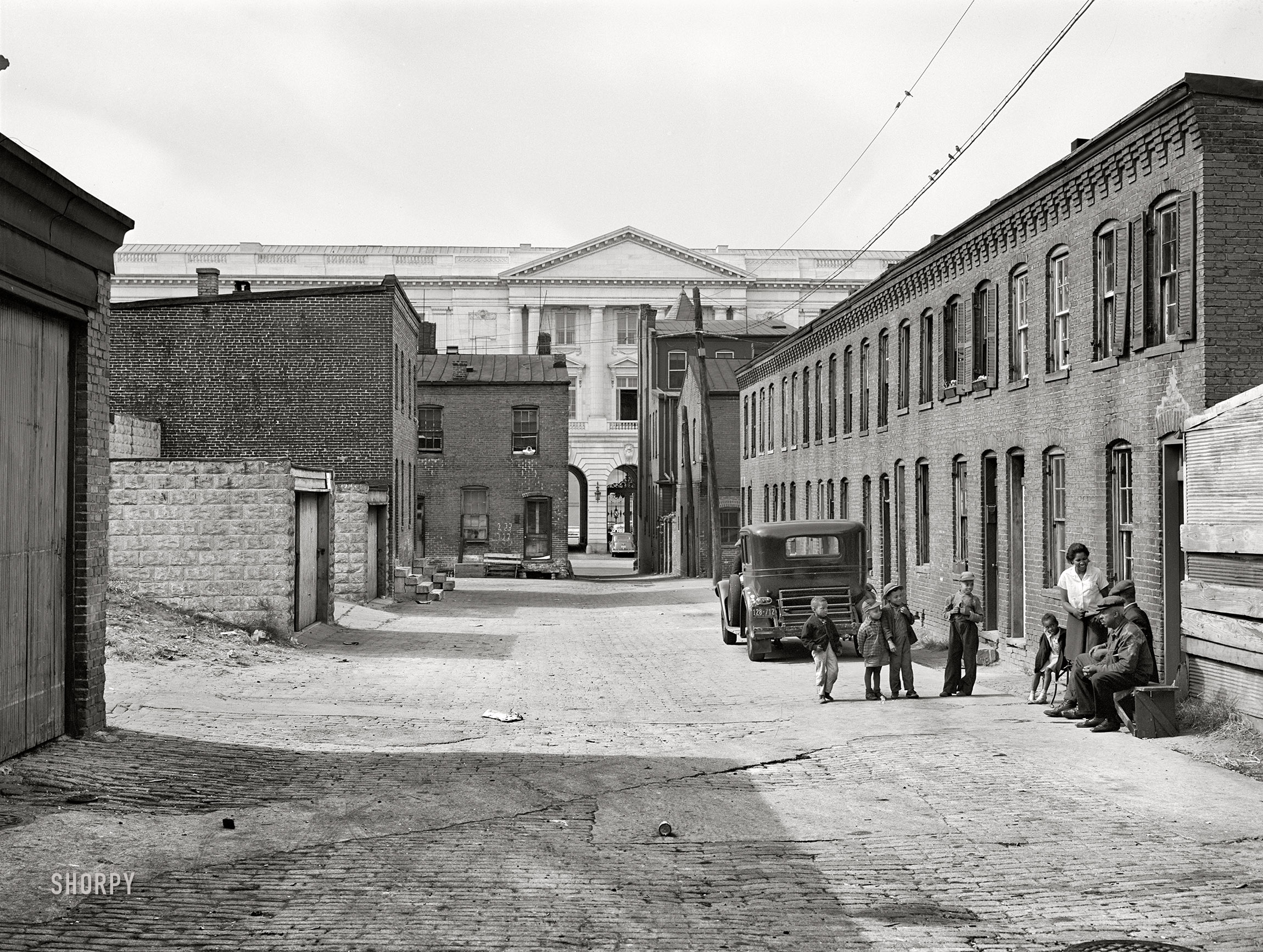 October 1941. "Schoot's Court [i.e., Schott’s Alley], Washington, D.C.; Senate Office Building is in the background. Four very small dark rooms rent for 15 and 18 dollars per month with water and privy in yard. It used to rent for 6 and 8 dollars. Frank Coles and his friend are sitting on the bench. He was a cement plasterer but has been on relief during the past year. He has frequent heart attacks and his feet and ankles are all swollen. Doctor advises a chicken and lamb diet, no pork or beef, but he doesn't even have money to buy fuel. He can't get waited on in a clinic or get to one. He waited from before 11 until 4 p.m. but still could not see a doctor. He has been in Washington since 1906." Photo by Marion Post Wolcott. View full size.