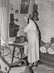 April 1940. "Lady signaling operator on old-style telephone. Scranton, Iowa." Medium format acetate negative by John Vachon for the Resettlement Administration. View full size.
&quot;Turn crank briskly ... &quot;In October of 1975 I drove my 1965 Falcon from Vancouver, Canada, to San Francisco on a leisurely trip down the coast. I arrived at a road junction north of Fort Bragg, and saw a phone booth. I planned to stay with friends in San Francisco, and thought this was a good time to call and let them know of my arrival day. Upon entering the booth, I discovered the pay phone had no dial, and below it was a black box with a crank on it. There were detailed hand-written instructions on how to use it. 
"Remove handset to be sure line is clear." There was no dial tone, and this was to determine if anyone else was using the party line. "Replace receiver, and turn crank briskly to signal operator." A male voice answered in Fort Bragg, and placed the call to San Francisco. The final instruction: "Turn crank briskly to clear the line". I charged the call to my Vancouver number, and when the bill came it read: "Call from North Rockport Toll Station No. 1." 
Vancouver was one of the last large cities to convert to dial phones, a 20 year process finally completed in 1960.
A visit to New Zealand in 1985 revealed that some small towns still had not received dial telephones, as shown in this photo of a pay phone in Taihape.
I have the wheat pastebut could you bring the brushes and seam roller when you come over to help hang the wallpaper today?   I have to burn some incense in the Buddha to cover up the smell from Bob's fish dinner.
Just When You ThinkIt could it get any better? Dave comes up with this caption!!! Now I have to add THIS ONE to my all time favourites list.
[Thank you! Although "Crank Call" is the title, not the caption. The caption is the writing under the photo. - Dave]
[Right! My old brain needs to reboot :) - Baxado ]
Roll CallShe has rolls of something in bundles and it seems she's calling the person she prepared them for to say they are ready.  Perhaps she is making some side money.  What are they? 
Corner shelfI made one in wood shop class identical to that one in junior high school around 1977.
Calling up CatfishMy grandfather (b. 1898) had the innards of one of those telephones in a bucket in his fishing boat. When he was hungry for catfish, he'd load me up and we'd go out to a deep bend on the Clear Fork of the Brazos and slide the jonboat through the reeds and into the river a couple hundred feet upstream of the bend.
On one of the copper telephone wires he had tied a horseshoe as a weight, which he dropped straight down off the side of the boat. The other, much longer wire was tied to a big iron washer -- about the size of a #10 can lid. I'd paddle down to the deepest hole in the river and he would swing the washer over his head like a lasso and toss it as far as he could. As soon as the washer hit the water, he'd holler "crank it!" and I'd wind the magneto as fast as I could, creating a current between the two poles and stunning any catfish caught in between them. After a few seconds he'd get the dipnet and scoop up any electroshocked fish and we'd repeat until he had enough for a fish fry.
I later discovered this was regarded as unsporting and possibly illegal means of harvesting fishflesh. In the mid-'60s, however, I never passed up and opportunity to go "call up some catfish".
Where&#039;s the bird?She looks a lot like Granny from the Tweetie and Sylvester cartoon.
Makes me think of Bryant Pond, MaineBryant Pond was the last community in the U.S. to have crank telephones, switching over to plain vanilla dial phones in 1983.
To call my friend in Bryant Pond, I had to dial 0 and ask the operator (remember operators?) to place a call to Bryant Pond 32 -- which was my friend's number. Some operators took it in stride; others took some convincing. 
Oh, my aching backI'm pretty sure her phone calls never lasted long - look at the posture she has to assume.  That table makes her stand about two feet away from the phone and then lean forward over the table to get close to the mouth piece.  I'm surprised I don't see a hand mark on the wallpaper from bracing oneself.  And of course she'd never just clear that table and sit on top of it.
She doesn&#039;t look BuddhistSeriously, I am a little surprised by the Buddha figure on her shelf.  Grandma was practicing way before it was hip!  
About those rollsI'm probably wrong but, those rolls seem to me to be too narrow. Is it possible that they are player piano rolls? A couple of them seem to have narrow wooden slats along one edge. I remember seeing player pianos in one of my aunt's homes when I was a kid in the early 1950s and they generally looked like these, but were usually stored in boxes.
No hands voice dialingThat phone used no hands voice dialing, an amazing technology that is virtually unavailable today.  
More!A person can never have too much wallpaper. Well, maybe sometimes.
 BuddhaMy grandmother had one -- an incense burner, in green-red-gold.
Isn&#039;t It Obvious?Those rolls next to her can't be anything but more wallpaper that needs hanging!
I really like the woodworking on that fern table in front of her! 
(Technology, The Gallery, John Vachon)