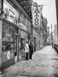March 1941. "West Main Street. Norfolk, Virginia." Medium format acetate negative by John Vachon for the Farm Security Administration. View full size.
Pick up your feetI'd bet more than one sailor on leave made a face plant on that sidewalk. 
Compact AfternoonA quick lunch, followed by getting that tattoo and a loan, then a haircut before dinner, all in the space of six storefronts.  Can't beat the efficiency!  
Hot DogI'd love to see that light lit up at night.
LOVE the Hot Dog SignThat sign tickles me to no end! In the words of the Three Stooges, Hot Dog, it's the Cat's Meow!
I&#039;d Swear That Guy In The Hatlooks just like The Kingfish!
One LeftThe only building that remains is the tall light gray one all the way down at the far right, past the tops of the parked cars. Everything else is long gone.  
Just a short walk from a long pierNothing identifiable in this picture remains today, aside from West Main Street itself. There wasn't much of West Main Street in Norfolk then, or now. Main Street becomes East Main Street when it reaches Granby in the next block or so, and behind Vachon was the end of West Main Street. Now, West Main Street leads to the Nauticus museum and entertainment complex and the pier where the U.S.S. Wisconsin is permanently docked.  
It&#039;s a Navy townSo I suspect that Coleman is not the community's only inker.
Now Hear ThisThree Light Cruisers approaching off the port bow.
All hands on deck and prepare to be boarded.
(The Gallery, John Vachon, Stores & Markets)