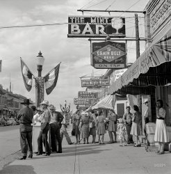 August 1941. "Main Street. Sheridan, Wyoming." An oasis of Western watering holes. Medium format negative by Marion Post Wolcott. View full size.
Oscar of Dutch LunchOscar Thielen of the Dutch Lunch.
Mint ConditionLove the bubbly-circles design on the dress of the woman with the white hat.  They're a perfect match for the "sun goggles" worn by the trooper a bit to her left.  
Oldest bar in Sheridan (1907)From a postcard (below, circa. 1945): "Renowned for its unique beauty and western atmosphere. Visitors never tire of the unusual red cedar decor; the western pictures and photographs; cowboy paraphernalia and the North America game animal trophies. Wayne W. Elkin and Leon L. McVean, Proprietors."
Musician Ben O’Connor on his visit in 2002: "The Mint Bar is the coolest thing about the great cowboy town of Sheridan, which is in a part of northern Wyoming that’s too cold and too remote for all the Californians to move there and ruin it. You wouldn’t want to say anything bad about John Wayne at the Mint. Real cowboys hang out there."
Old school coolPretty fly for a cowboy guy.
hoooo-EEEEEThe riding-heel boots on them two ol' cowpokes to the left ... they's wearin' more heels than the women are!
Indiana Jones takes a vacationNo hat, no coat, no whip; just hanging out in front of The Mint chatting up a pretty lady.
Still thereLocated at 151 Main Street (US 14). The buildings across the street are intact, but the shops on the right have been replaced by "storefront" school -- Sheridan College (in what appears to have been a former department store).
Going DutchI wonder if "Dutch lunch" meant the same thing there as it did in the Detroit of my grandparents' era - a lunch taken with a beer (or two)?  In fact, sometimes the "Dutch lunch" could be just the beer, no lunch.  My grandfather, though Irish, was certainly not adverse to enjoying an occasional "Dutch lunch."
Lady on the far rightI'm sure I brought the baby...
Re: Old school coolIt's not quite contemporary, but I remember just such round-lensed heavy-framed sunglasses from around 1950 as having two polarized lenses over each eye so that one lens could be rotated to vary the amount of light getting through.
The Cowboy Named &quot;Doris&quot;The straw-hatted cowpoke in profile, left of frame, with his hand resting on the letterbox looks a lot like an old cowboy I chanced to meet over a game of “8 Ball” in a little bar in New Mexico four decades to the month from this photo’s date.
We played a couple games swapping dimes on the table while he smoked hand-rolled cigarettes hanging from the corner of his mouth just like the guy in this photo. After setting me up by letting me win one we bet a beer on the next game and as he was running the table on me, calling pockets by gesturing with the tip of his cue or a nod or his elbow when indicating a long-green, corner pocket shot banked off first the long then the short rail and all the way back down the baize to the corner at his right wing, I introduced myself and he said his name was “Doris”.
I though I might have misheard and used his name when I bought him his Coors and he answered to “Doris,” thanking me for the beer with a touch of his hatbrim and squatted to retrieve the balls from the slot near the jukebox end of the table and put them in the triangle and returned it to the hook below the ball return. 
Doris took a couple long draws from his beer and wiped the can’s condensation from his hands onto his shirtfront and took out papers and tobacco from his left breast pocket (just like the photo cowboy) and rolled another smoke and I asked him how he got the name “Doris” and he said he was the seventh of nine children and his parents ran out of names at about number six. "After that they wrote some names on little slips of paper and drew them from a hat when babies were born," He said as he struck a big wood kitchen match on the coin slot of the pool table, “If you think 'Doris' is funny, you ought to meet my sister '6 7/8'".
Goober Pea
Well it ain&#039;t changed muchAnd that's the way we like it around here. 

The glassesThis image is plastered on a poster around town here in Montreal, advertising an exhibition at the McCord Museum of the photographs of Horst P. Horst (1906-1999).  The photo is of Muriel Maxwell and was featured on the cover of American Vogue on July 1, 1939.
Sheridan College nr Mint Sheridan College is in the old Woolworth's building, across the alley from the historic Mint Bar.
About 1990, Queen Elizabeth II visited Sheridan for the horses. She shopped in a fishing-gear store, next door to the Mint. It was said that she visited the Mint, but I never believed it. Instead, she crossed Main and stopped in the King Ropes store for a visit to the renowned rope, tack and saddle emporium. Her host was WY Sen. Malcom Wallup. 
Them&#039;s Ridin&#039; JeansNote the back pockets on the jeans on the cowboy all the way to the left, in the foreground (black hat). They are set way off to the sides, towards the hips, unlike today's jeans with pockets near the rear-end mid-line. Wouldn't want to be riding with your wallet between you and the saddle.
(The Gallery, Eateries & Bars, M.P. Wolcott)