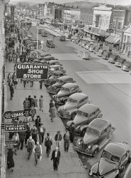 December 1940. "Christmas shopping crowds. Gadsden, Alabama." Medium format acetate negative by John Vachon for the Farm Security Administration. View full size.
Fur realWoman wearing fur #1 (her head is immediately to the right of Rains &amp; Rains Lawyers) is turning to check out woman wearing fur #2 (just above Use Stair Way).  The real challenge (aside from identify all the cars) is: identify fur #2.
Dan Cohen is selling Hoes?!And on Broad Street, no less.
I want to respond to sinking_ship and his "Bag the drama" comment. Yes, there are possibilities as to what exchange is taking place; but there is also probability. Most who have commented believe the black man is probably being insulted.  And just as he is not looking at his white antagonist,  the antagonist's friend is also looking straight ahead, without expression.  I read that to mean he wants no part of what his friend is doing.  And you ask does any of this affect you? -- it does, because all of us today need to accept and acknowledge that we cannot treat minorities the way our grandpas did.
Why are you looking at these old photographs if you feel no compassion for the subject matter?
Can&#039;t see Gadsden for the treesHere's the approximate place - it's hard to see because of the trees planted sometime after 1940. Sterchi's Furniture was at Fifth and Broad, and we can see the Belk-Hudson (Hughes 1903) building with the curved windows on the right (all white in the original).

The South in the 1940sThe first thing I noticed was in the foreground, the fellow in the coveralls with his back to us.  The look the guy on his right is giving him ...  To me, it's very chilling.
Snark?In the lower left corner, there is a White man who appears to be making a comment to Black man as they pass each other. One wonders about what is being said, in 1940's Jim Crow Alabama. 
Fur in AlabamaSure doesn't look cold enough for fur, based on the dress of everyone else.
A classic lookThat guy in the fedora near the bottom of the photo, second in from the curb, is displaying one of my favorite "looks" from that era, with the leather jacket and the khakis and the hat. That will never not look "cool." 
CarchitectureInteresting how much cars of the same vintage -- no matter the make -- tend to look so similar.
VisitorMost of the cars seem to have the proper Alabama plates for the time, but the third car up from the bottom is an out-of-state car, though I do not know from where.
Here and now...There are so many other possibilities as to who or what caused the gentleman to turn his head. Should we dox his descendants and make sure they pay for his vile transgression that is so clearly explained from a shutter click 80 years ago. Bag the drama, please. 
Sneak peekThe briskly walking elegant lady, bottom left, in the dark coat has what can only be described as a well-turned ankle ... and the overalls-clad gent with the package under his arm might have noticed it. Also Rains &amp; Rains lawyers ... that's pretty funny. Must've been plaintiff attorneys, perhaps collaborating on larger cases with Monin &amp; Gronin, Attorneys at Law.
New legal partner?I was waiting for one of the Rains legal partners to leave, and a new partner to come on board: Pours.
Albert McKinley Rains (1902-1991)Rains was born in Grove Oak, DeKalb County, on March 11, 1902, to Elbert and Luella Rains; he had three siblings. He attended local schools and after graduating high school attended John H. Snead Seminary in Boaz, Marshall County. Rains went on to attend present-day Jacksonville State University and the University of Alabama. He studied law, passed the bar exam in 1928, and practiced alongside his brother Will Rains at the firm of Rains and Rains in Gadsden the following year.
http://encyclopediaofalabama.org/article/h-3683
Re: Fur in Alabama? Being from the South in my youth, until I married at 19 and moved North with my new husband at 20 when he got out of the Air Force, and growing up around plenty of older, well-to-do Southern Ladies, I seem to recall that the biggest rule of proper dress had little to do with the temperature, as long as the season was right! 
As long as it's not hot enough to cause a lady to "glow" - gentlemen "perspired," and horses would "sweat" - then a properly dressed upper class lady, (providing she owned one, which one couldn't be a proper lady without one) wouldn't be seen in public without her fur coat, (and matching or coordinating hat!)
Visitor: A Yankee!The third car up from the bottom highlighted in a previous comment displays a 1940 Massachusetts plate.  And wouldn't it satisfy a bit of curiosity to discover the plate's owner in the MA registry of motor vehicles (assuming the archives go that far back)?
Not only todayThey say most cars today look basically the same. I guess that could apply to most of the cars in this photo too. 
(The Gallery, Cars, Trucks, Buses, Christmas, John Vachon)