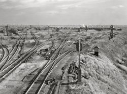 November 1942. "Chicago, Illinois. General view of the north classification yard at an Illinois Central railyard." Acetate negative by Jack Delano, Office of War Information. View full size.
All downhill from hereThis image shows the many power switches and retarders used to direct and control the speed of cars that have been shoved over the top of the hump and roll by gravity to their intended classification tracks.  The hump yardmaster (located behind the photographer) arranges the switches and retarder operators (in their small towers) slow the cars by squeezing their wheels so that they roll their intended distance before coupling up to cars already in the track.  Wind, different car weights and number of cars already in the track require a lot of judgment to prevent a car from "stalling" before reaching its intended destination or rolling too fast and slamming into a standing car.
I count 13 guysThe north classification yard is not as abandoned as first appears.  There are two men in the center foreground, huddled over working on something together.  Then, straight up from them and a little to the right, is a man walking the tracks.  Beyond him, where the railroad cars are, I count 10 men walking (I'm pretty sure they're all men).
I hoped to see one of the control (switch?) towers occupied, but no.
(The Gallery, Chicago, Jack Delano, Landscapes, Railroads)