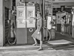 December 1942. "New York, New York. Girl at gasoline pump." Medium format acetate negative by Royden J. Dixon for the Office of War Information. View full size.
Outfit materialVelour?  Corduroy?  It has a soft, plush look.
8 cents per gallon?Am I reading the price correctly on the gas pump?
[Um, no. - Dave]

Fill &#039;er Up!Tell me that's not Ethyl manning the pumps!
Neatness countsWomen employees had certainly improved the look of stations since John Vachon photographed one two years earlier.
The smoking sectionA fine example of modern looking "computer" pumps, at least if we ignore the archaic sight-glass (and those weird dome tops, that give them the appearance of small mosques).
What particularly caught my eye, however is the curious(ly skimpy) "No Smoking" signage: the placement gives the illusion that the proscription only applies to the regular grade  
That is no girl!An adult woman, to my eye. But those were different days.
&quot;Extra&quot; pumpsWonder why there are two "extra" pumps sitting inside the station in the background apparently not (yet) in use.  Maybe the station was in the process of converting over to the "new" style pumps and that's what motivated this photo.
[There are pumps on both sides of the island. What motivated the photo is wartime girl station attendants! - Dave]
Price per gallon todayAdjusted for inflation, 20 cents in 1942 would be $3.83 today. 
Outfit material continuedI guessed it might be cotton velveteen?
Want to feel old?Few people who are less than solidly middle aged have seen gasoline pumps that show a sale's cost with only three digits.   Yet before the 1973 oil embargo they were ubiquitous.
PeekabooI wonder who is that hiding behind the bulk oil dispenser? Doesn't appear to be wearing a pump jockey uniform.
When People Didn&#039;t Throw Away Pennies8 and 10 cents a gallon are $1.56 and $1.95 in today's dollars.
[The prices on these pumps are 18 and 20 cents a gallon. - Dave]
Mystery equipmentWhat is the equipment on the right in front of the two pumps and the person in black on the back side of the island?
Gas Is Less Expensive Today If 20 cents equals $3.83 today, one must remember that cars today get almost twice the mileage they got back then. So back then to go to and from the same distance as one would go today it would cost them 40 cents, adjusted for inflation should mean that gas today should be $7.66.
IMO, the reason we think gas prices are so high is because of all the "stuff" we simply must have that did not exist back then, like computers, cable TV, the Internet and more, that comes out of your paycheck each month. Take all that away and $3.50 a gallon would be less a drain today than 20 cents was back then. 
In Case of Fire ... There is a handy soda-acid fire extinguisher hanging on the wall to the left. It appears that there was some wear and tear on the filler hoses dragging on the ground. The fix was to wind some heavy rubber tubing around the area on each hose. The Imperial Oil Company in Canada still uses the name Esso for its gas stations. 
Silver LiningCoins were made from real silver back then!  Take that into consideration when adjusting for inflation.
(The Gallery, Gas Stations, NYC)