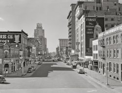 November 1942. "Oklahoma City, Oklahoma -- Hotels on West Grand Avenue." Medium format acetate negative by John Vachon for the Office of War Information.  View full size.
Urban RenewalOn the right, the building with the "Fidelity" sign was known as the Baum Building. It was one of the most ornate and regretted demolitions of the 1970s "renewal". I have a small piece of it. Just beyond it is the Colcord Building, the taller white structure, which is the only thing in this photo that remains. Built as office space in 1910, now a high end hotel. https://www.colcordhotel.com/about  Next is the Warner Theater located in part of the site of the current Devon Tower, tallest building in the State. Just beyond that is the Black Hotel and the Union Bus Station. Both survived until about ten years ago.
Across on the left is the 28 story Biltmore Hotel. For many years it was the largest structure brought down by implosion. I witnessed that one. Now part of the Botanical park mentioned in a comment on Seed Town.
Grand has since been renamed Sheridan.
Warner TheaterI see the Warner Theater in the distance. My dad worked there in 1946/1947.
Renewed
Just before the Warneris a tall white building. It is the only building in the picture that still exists, and is now an upscale hotel. Where the Warner Theater stood, is now an 845-foot-tall building, home to an energy company. I once saw "This Is Cinerama" at the Warner in about 1957.
Park-O-MeterCarl Magee of Oklahoma City  invented the parking meter. See his great creation at its birth above. In a way he helped the rise of the mall with its free parking and the demise of main street. What a legacy!
Ka-BOOM! townNote the Biltmore down the street, a prominent example of a celebrity implosion (right around the time when they became popular as new stories and cities began to search for some prominent, hapless building to be "honored")

And to build - no pun intended - on 'Studebaker1913's post: another hapless building (tho not imploded)

Among its sins: "I.M. Pei wanted to clear the Venetian Style Baum Building in order to straighten Robinson Avenue." Oh, my.
Being HumansOnce again the startling and heartbreaking contrast between the past and the present. Then; a street for human beings. Park where you want, walk where you like. Get a meal, buy a drink, find a room, hock your saxophone, maybe do a little shopping. Be human. Meet other humans doing human stuff. Now; some kind of corporate hell. Nothing to do, nothing to see - drive right through.
And how is it that, once again, a black and white photograph looks sunnier and warmer than Google street view?
Why so many hotels?Since some of the commenters have personal, historic knowledge of OK City, I'll ask: why are there so many hotels along this stretch of West Grand?  I found there were two railroad stations a block or so behind where John Vachon was standing.  The Santa Fe station is still there; the Missouri–Kansas–Texas station on East Reno is gone.  You can spin the Street View provided by Studebaker1913 around to see the train overpass.  The Santa Fe station is to the right.  Was there something else in this area to make so many people want to bed down nearby?
Today, there are fewer, but much bigger hotels.  On the immediate right in Street View there is a Wyndham and a Sheraton.  Down the street is the aforementioned Colcord.  But I figure they're here because, on the left in Street View is a convention center and then a sports arena on the other side of Reno Avenue.
I&#039;ve got my dancing shoes on, but my wallet is in the carThe Tap Room has "free dancing" but charges for parking. I guess they know a good racket to run!
The True Inventors of the Parking MeterWhile Carl Magee had the initial idea for the parking meter and he received a patent for it in 1932, he was unable to make a practical working model until he enlisted the help of two engineering professors at Oklahoma Agricultural &amp; Mechanical College (now Oklahoma State University, my employer) in Stillwater, Oklahoma. They were H. G. Thuesen and Gerald Hale, who perfected the design in 1933. The first batch of 175 parking meters was installed in downtown Oklahoma City on July 16, 1935.
Urban Renewal - UGH!Yet another Shorpy photo depicting an American city or town of yesteryear that looks so much better than its modern counterpart in Google Street View.
Why Hotels?In response to Doug Floor Plan, I would speculate that this phenomenon was quite common in most US cities in 1942.
Today's hotels are scattered throughout metro areas, especially at freeway interchanges. At that time, there were cabin/cottage like motels out on the highways, but hotels were almost always centrally located. In this case, there was even a third rail station (Rock Island) located a few blocks south to further increase traffic.
By the 1960s, places like Holiday Inn were showing up on the highways, blurring the lines between Hotel-Motel.
For another survivor of the old hotels, navigate two blocks north at the first intersection on my street view link to see the Hilton Skirvin on NE corner of Park &amp; Broadway. We almost lost that one several times. Prior to 1933 the Rock Island station was located directly behind it. The tracks were relocated south to avoid having east-west lines running right through downtown. Many City and County Buildings were developed in the mid 1930s along the former line.
(The Gallery, Cars, Trucks, Buses, John Vachon, OKC)