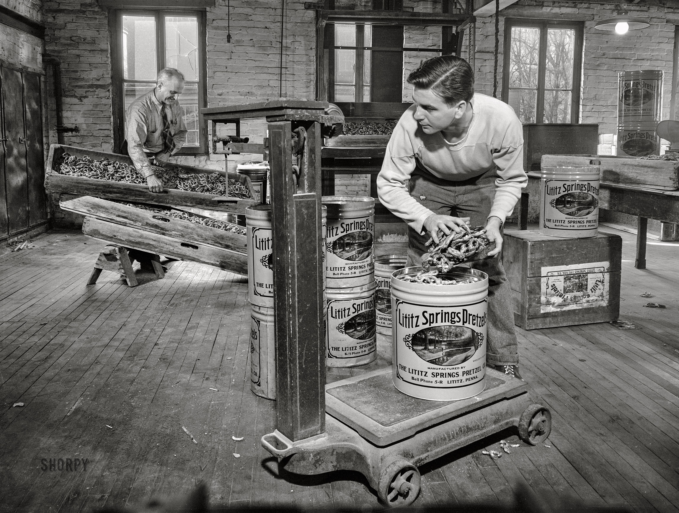 November 1942. Lititz, Pennsylvania. "Lititz Springs Pretzel Company, owned by Lewis C. Haines (background), who is unloading a tray of pretzels which has come up on a dumbwaiter from the baking room below. A son, Bob, weighs them and packs them in cans. Lititz was the first town in America where pretzels were made." Photo by Marjory Collins. View full size.
